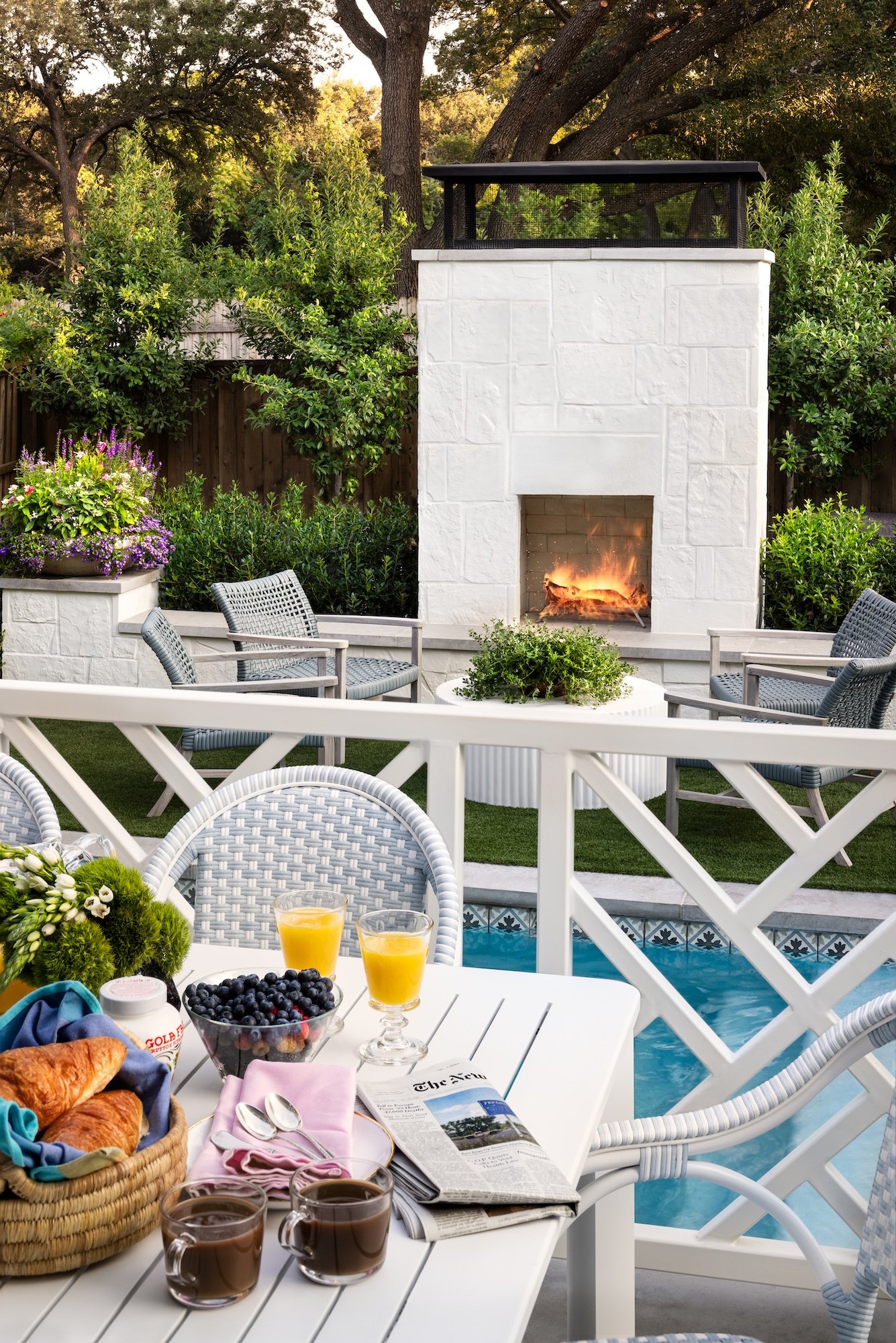 An outdoor patio breakfast scene with a white table and chairs, breakfast foods including croissants, blueberries, orange juice, coffee, and a newspaper, a fireplace in the background, lush greenery, flowering plants, and a swimming pool.