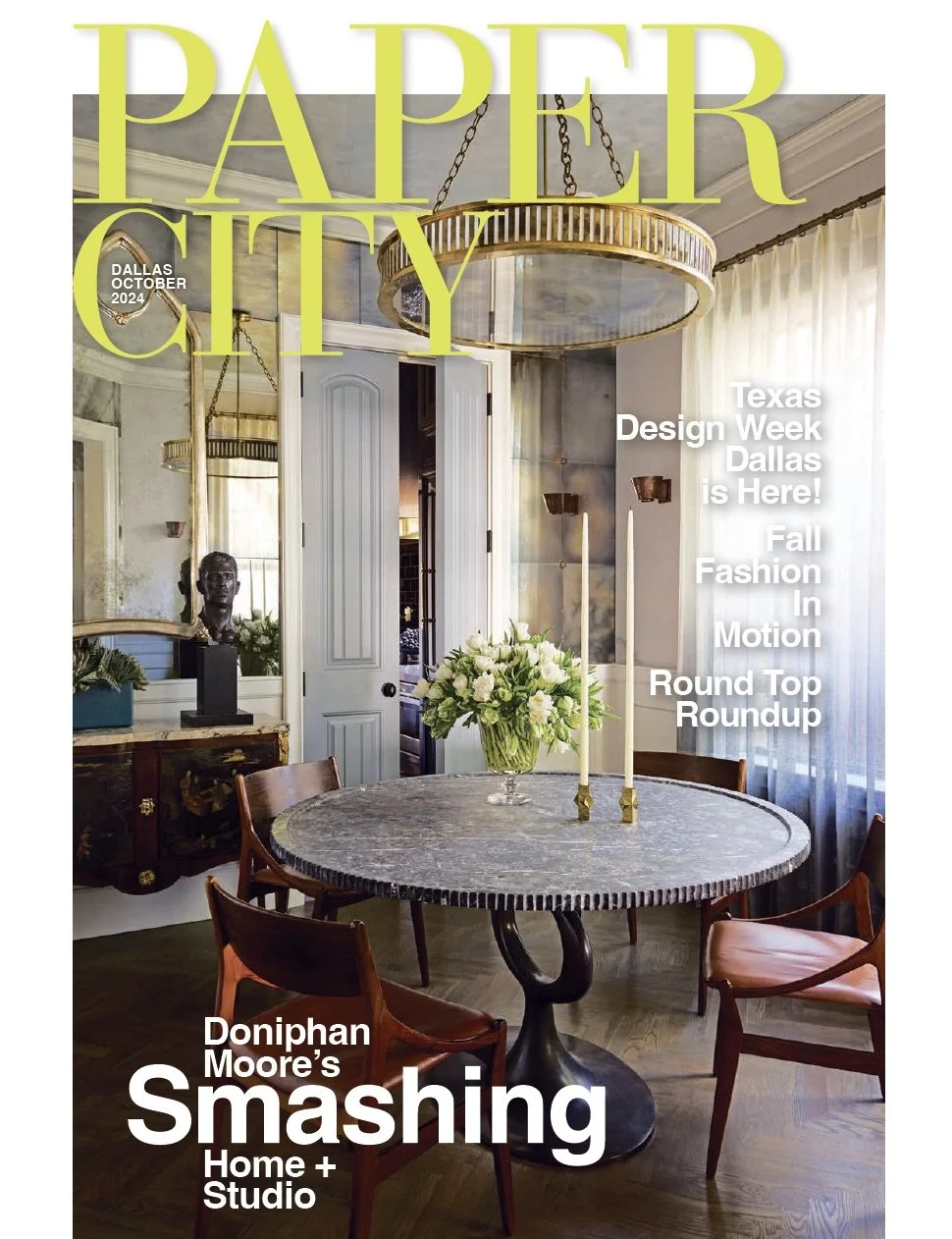 Cover of Paper City magazine featuring a dining room with a round black table, wooden chairs, floral centerpiece, and large windows with sheer curtains.