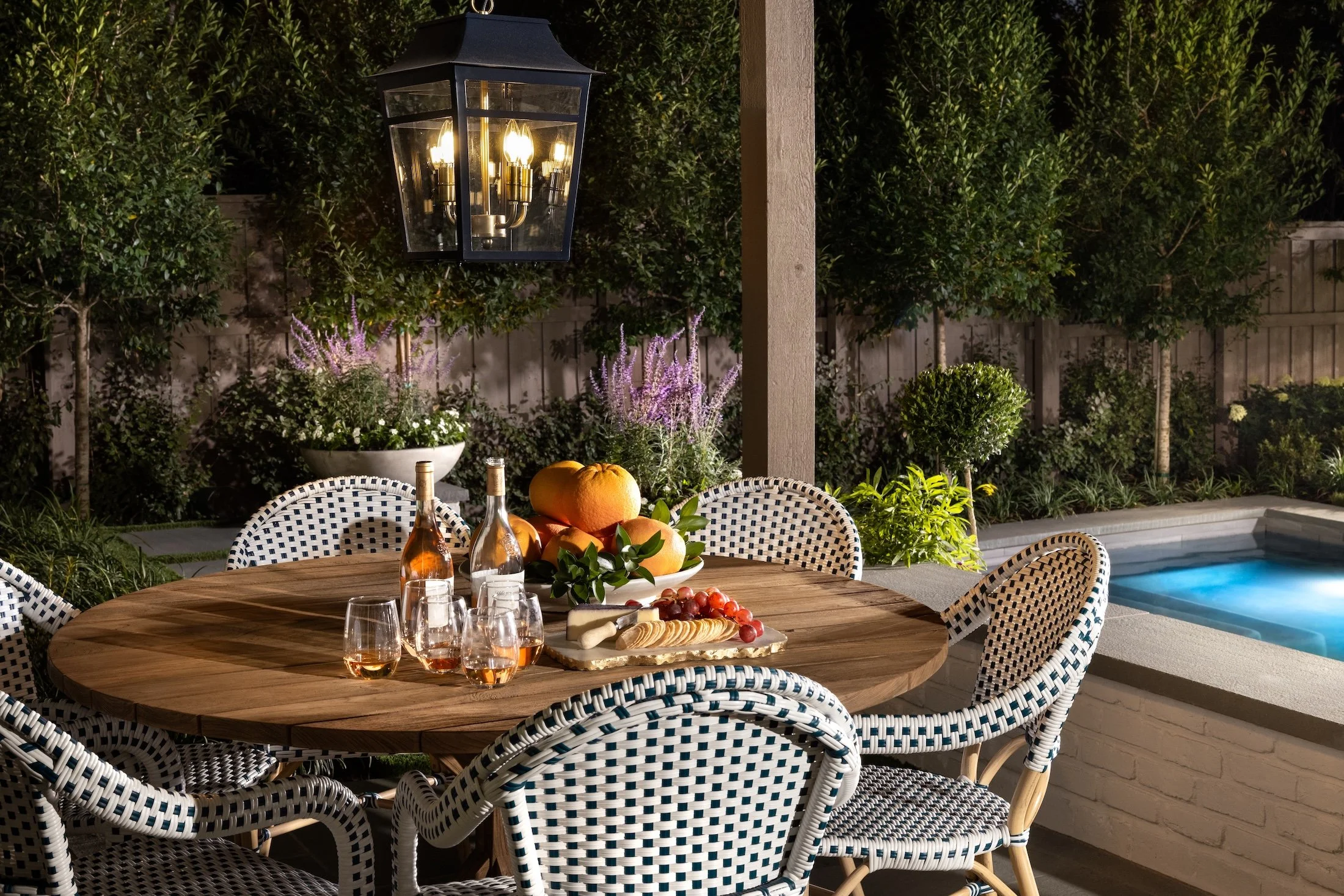 Outdoor dining table with drinks and snacks next to a pool at night, illuminated by a lantern-style light, with trees and plants in the background.