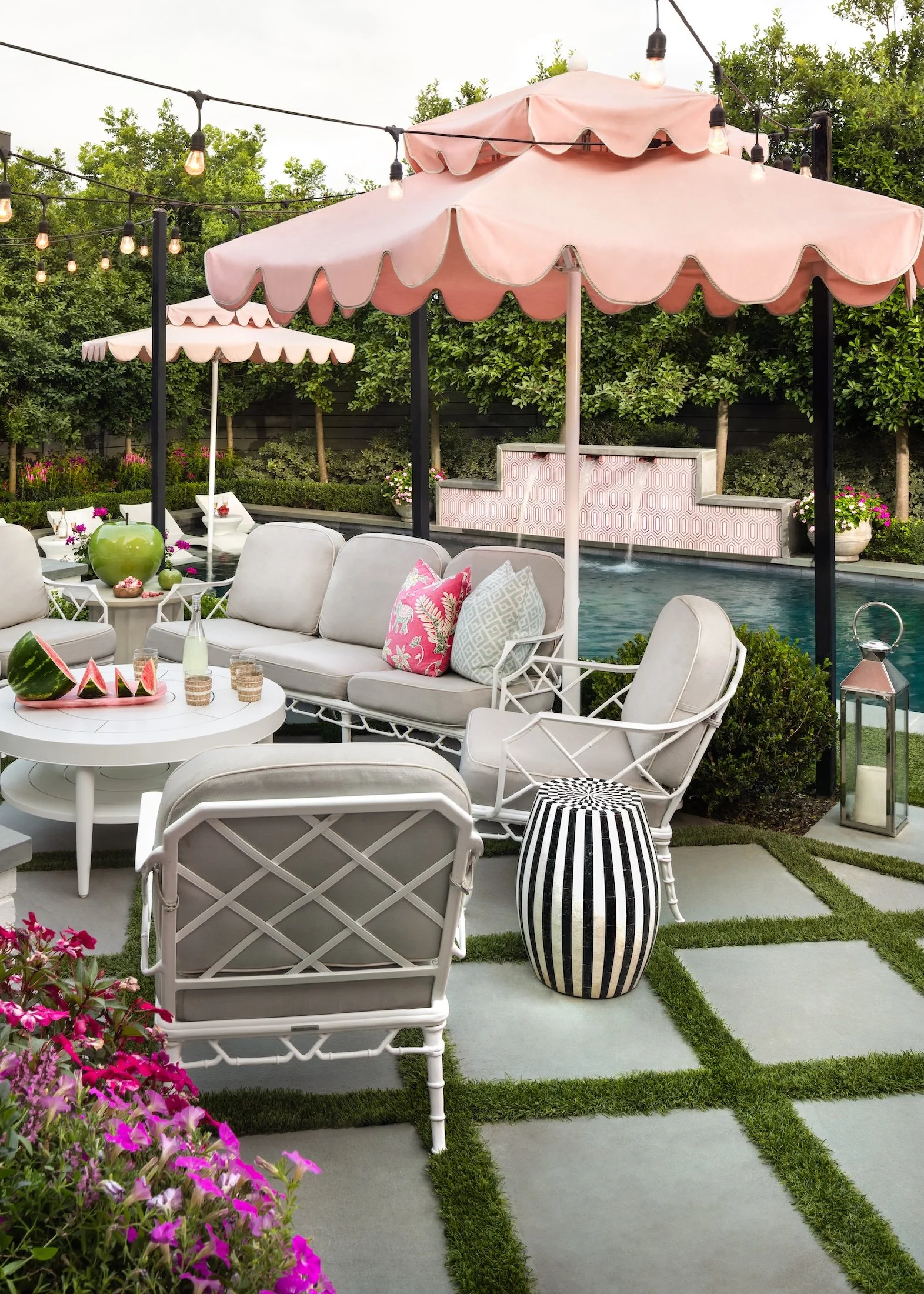 Outdoor patio area with white cushioned seating, pink umbrellas, and string lights, next to a pool and surrounded by greenery and pink flowers.