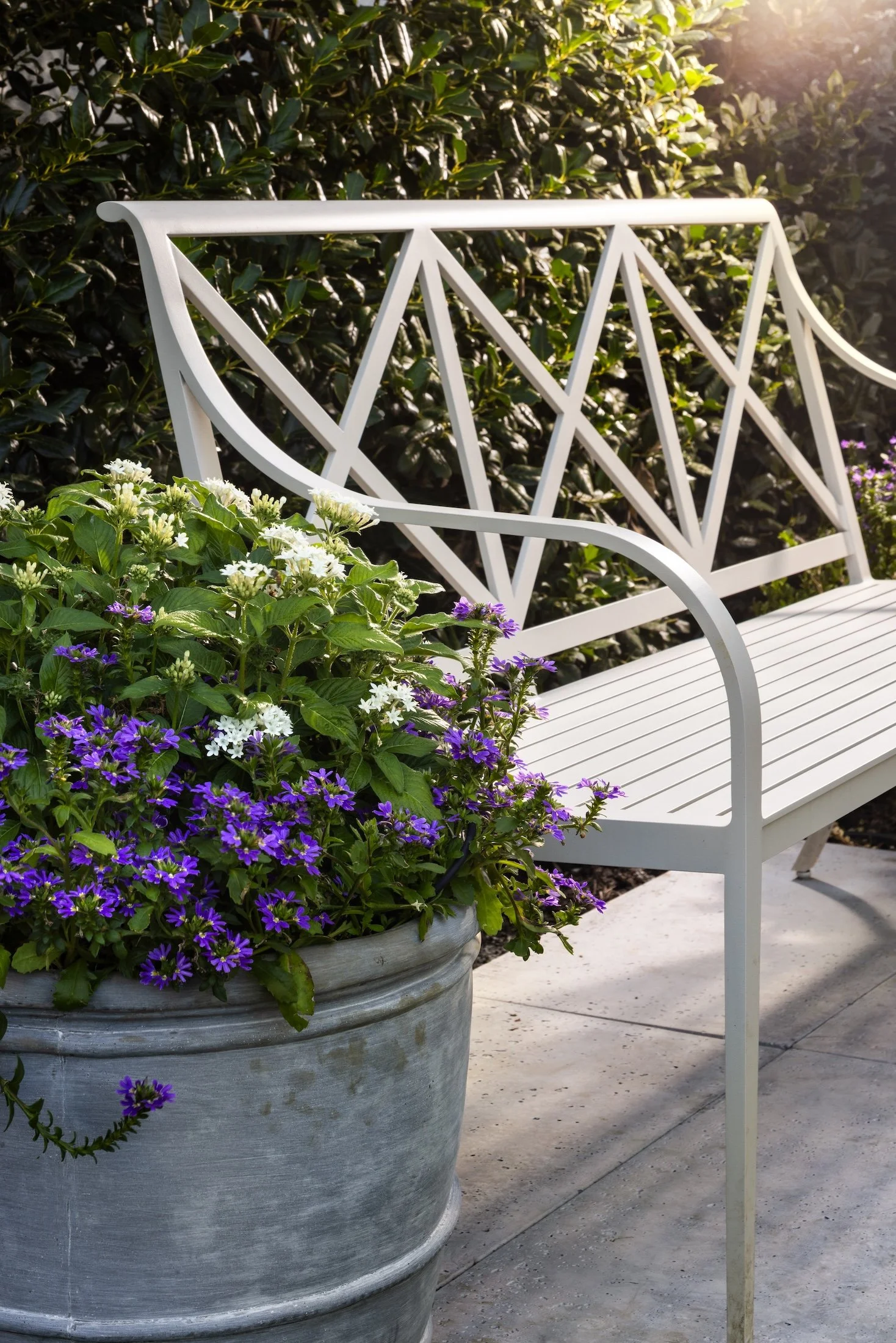 A white metal garden bench situated next to a large pot of purple and white flowers, with greenery in the background and sunlight shining from the upper right corner.