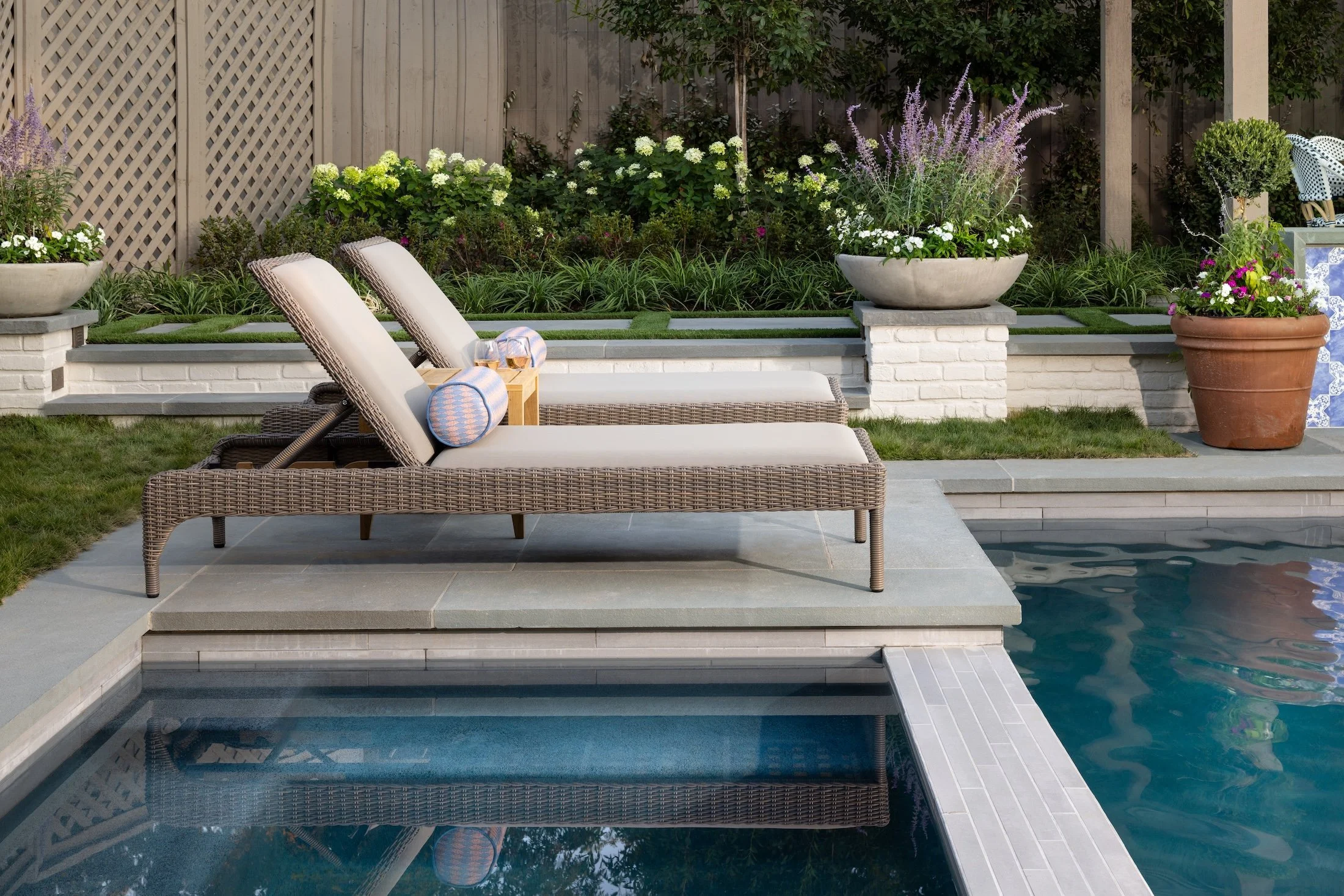 A backyard with a swimming pool, lounge chairs with pillows, potted flowers, green grass, and a wooden fence with lush plants and trees.
