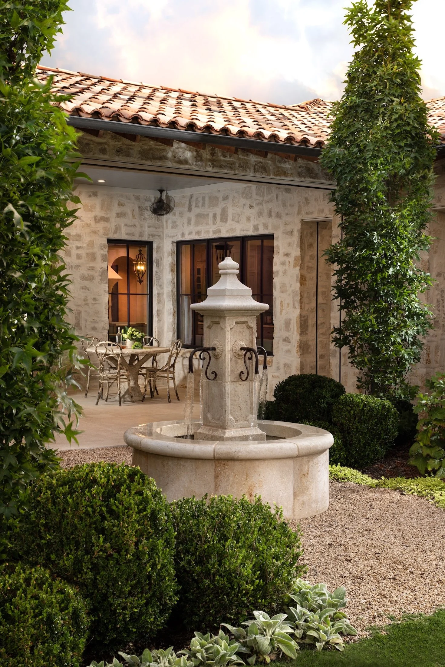 A stone fountain in a lush garden courtyard with green shrubs and foliage, a round patio table with chairs, and a house with stone walls, windows, and a tiled roof in the background.