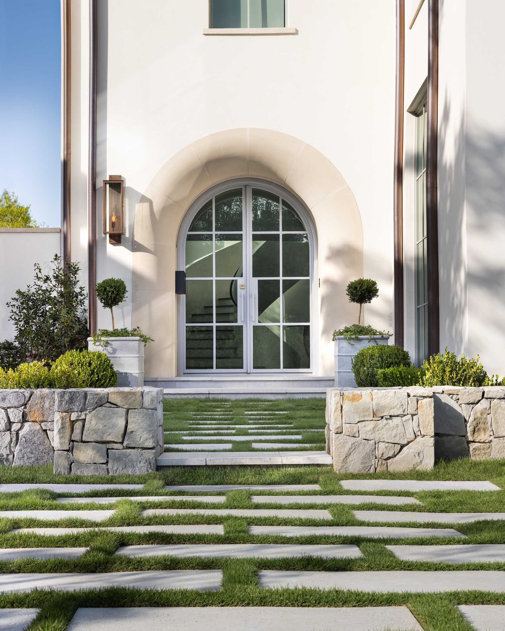 Front entrance of a modern house with a glass door, stone pathway, and landscaped garden with bushes, trees, and a stone border.