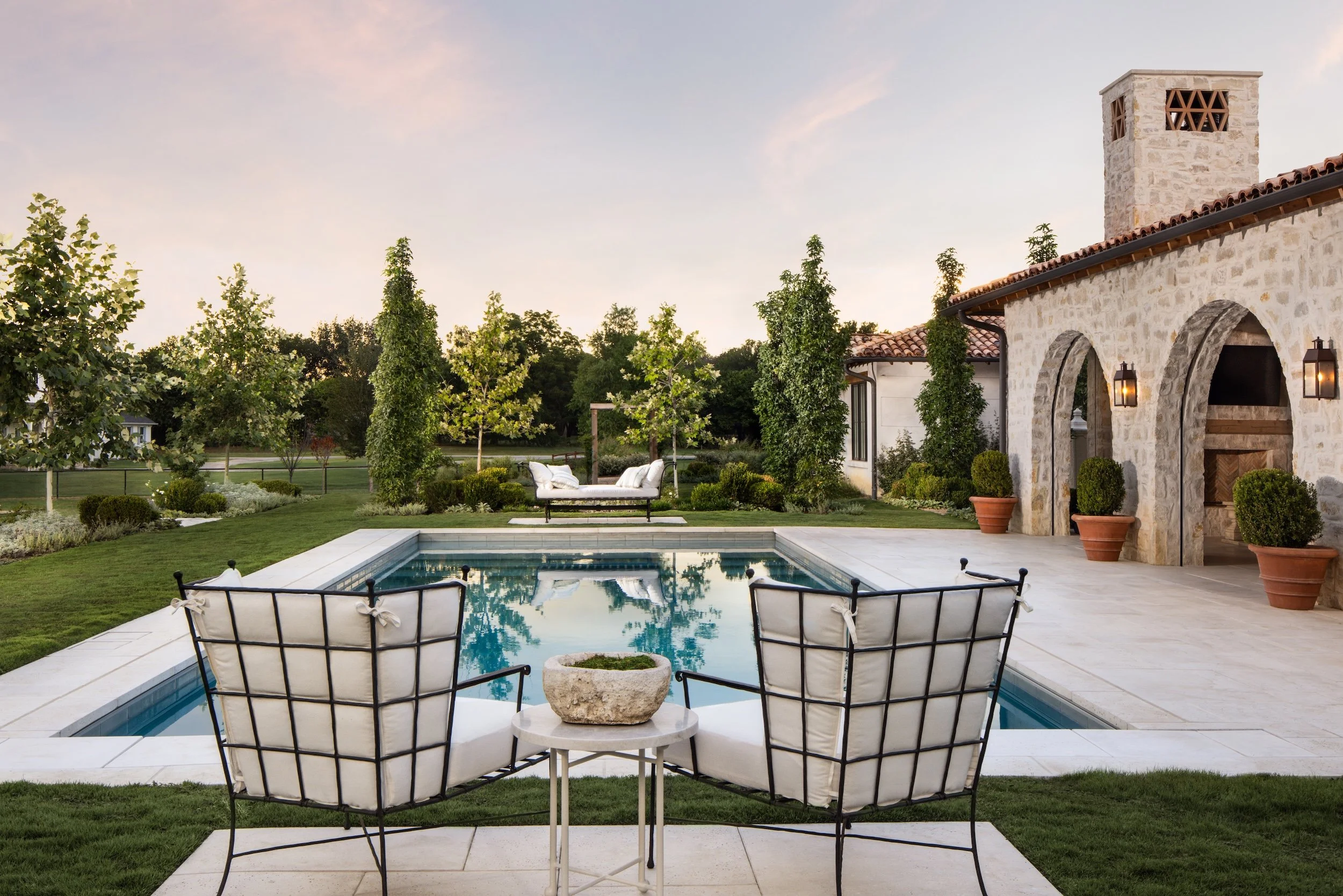 A backyard with a swimming pool, surrounded by greenery and outdoor furniture, and a stone house with a tower.