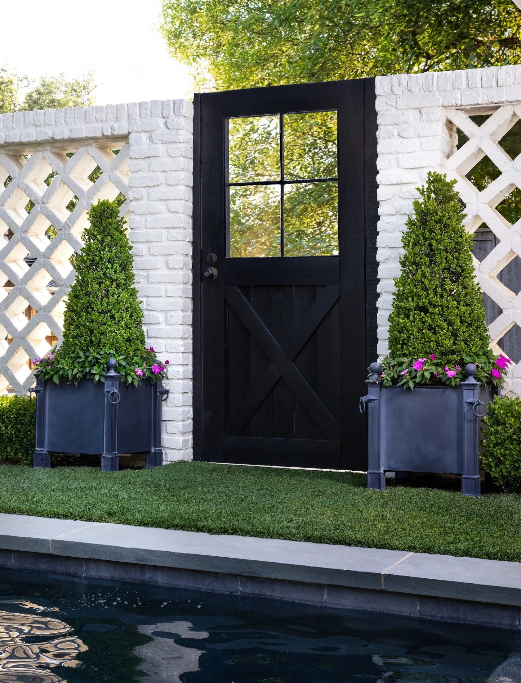 A black wooden gate with a window, set against a white brick wall. Two planters with green shrubs and pink flowers on either side of the gate, on a grassy area near a pond with reflected trees.