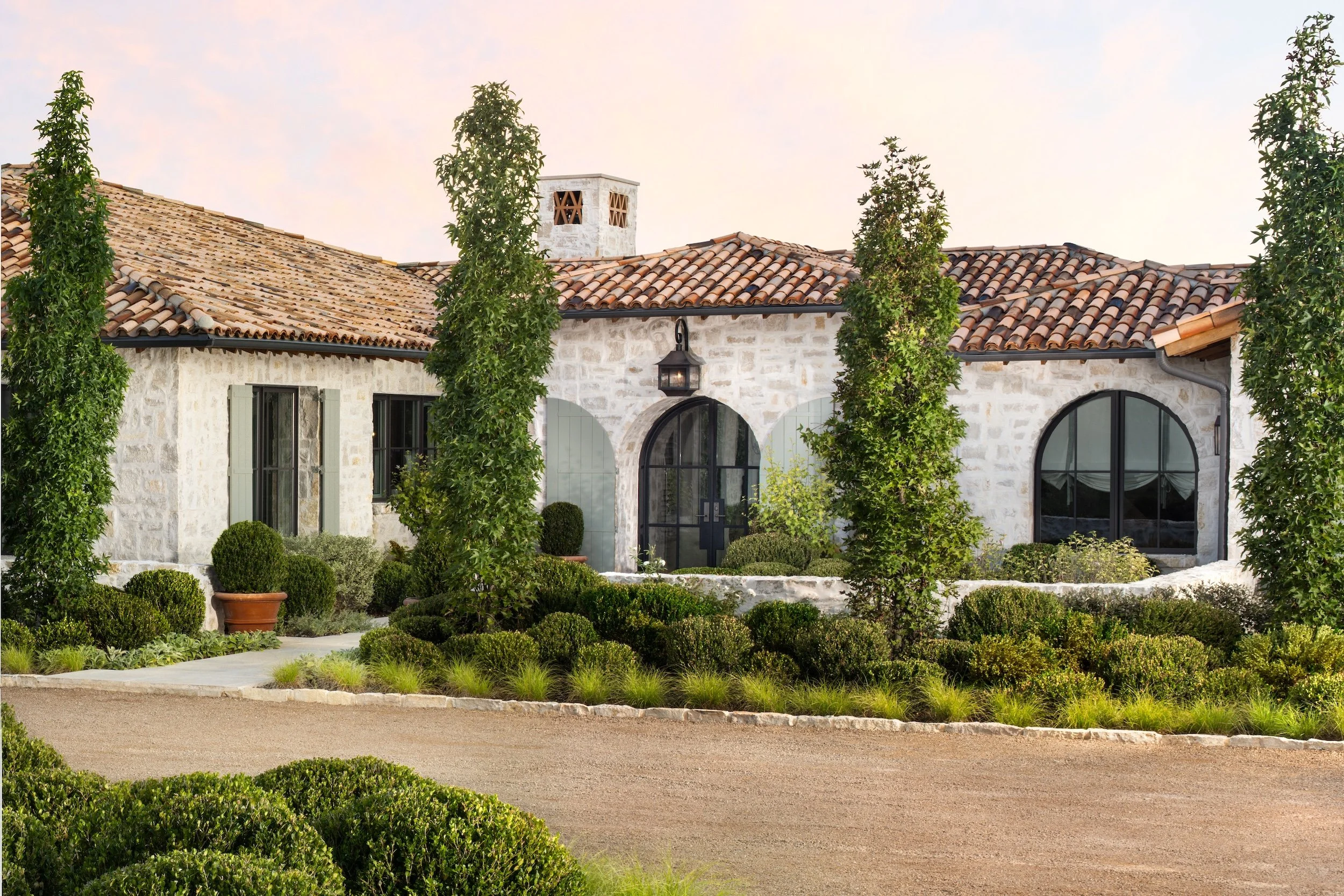 A house with white stone walls and a red tile roof, surrounded by manicured bushes and trees. There is a large arched glass door and a lantern-style light fixture above it. The sky is clear with a light pastel hue.