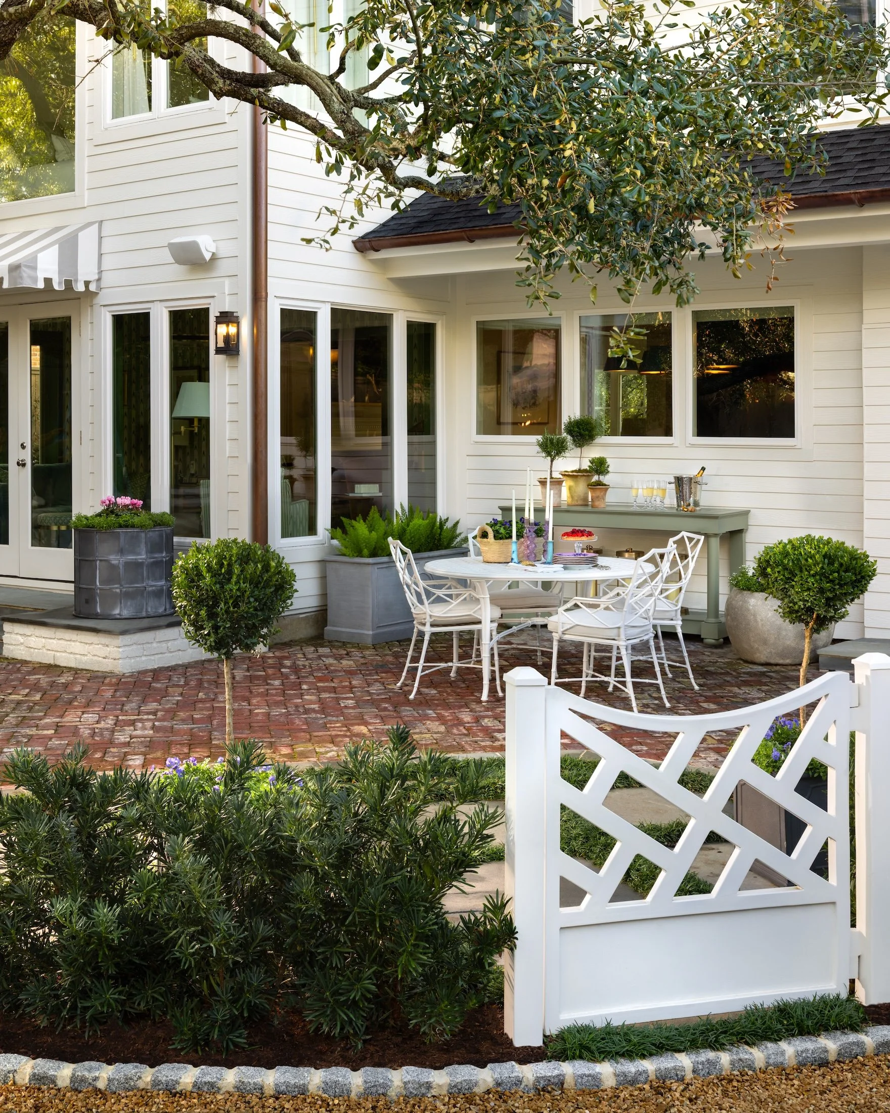 A white house with large windows, a patio area with outdoor dining furniture, potted plants, and decorative candles. A white lattice fence surrounds the garden with greenery and small trees planted in the yard.
