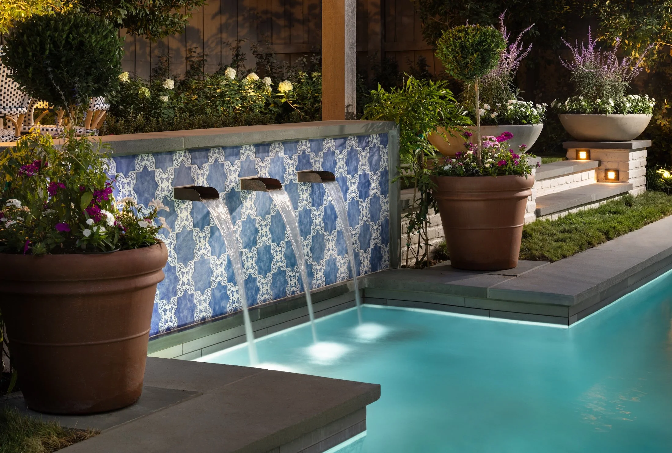 Nighttime scene of a backyard pool area with potted plants and illuminated steps, featuring a water fountain with water flowing from spouts under a tiled decorative panel.