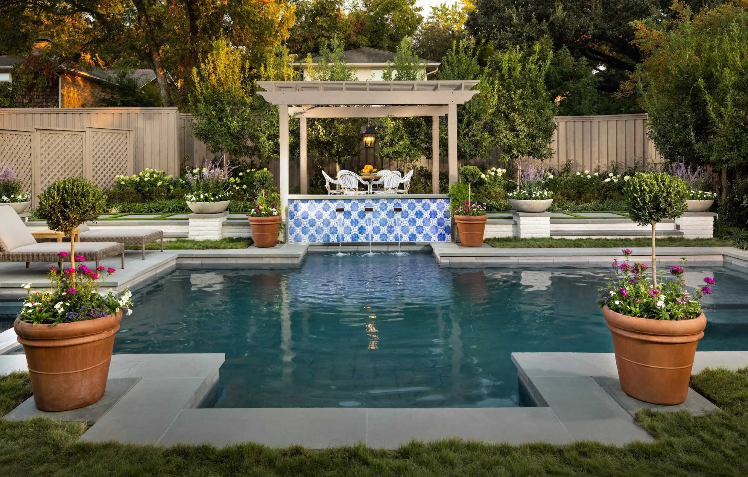 A backyard with a swimming pool, surrounded by potted plants, trees, and a white fence. There is a covered patio area with a table and white chairs, and a decorative blue tile fountain with water flowing into the pool.