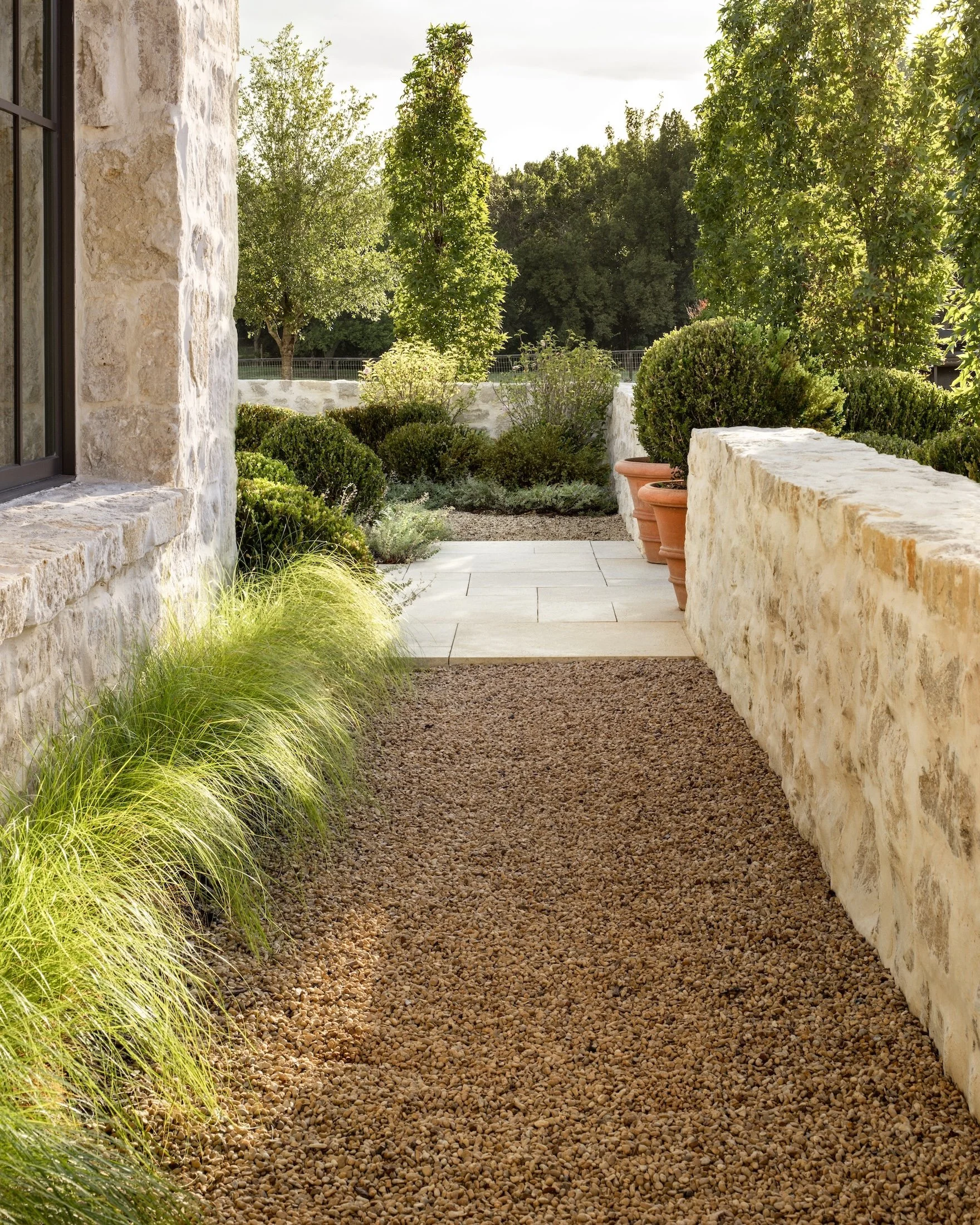 A stone pathway bordered by landscaping including grass, bushes, and potted plants, leading to a garden with trees and a fence in the background.