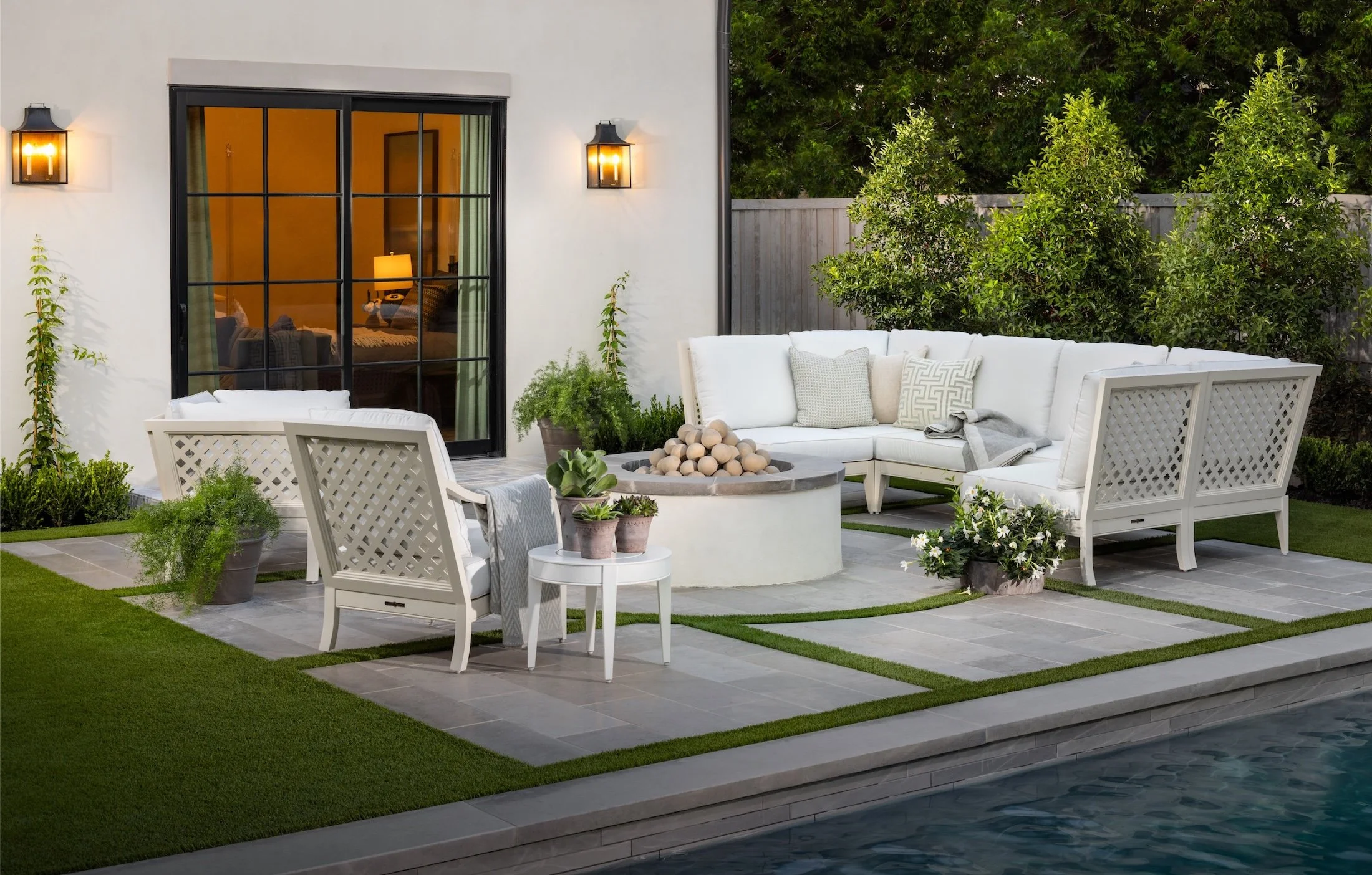 A modern backyard patio at dusk with outdoor furniture, including a white L-shaped sofa with decorative pillows, a matching armchair, and a small white side table with potted plants. There is a fire pit with decorative stones in the center, surrounded by potted plants and small green patches of grass. The house features large glass doors with black frames, and outdoor wall sconces provide warm lighting. A wooden fence and lush green trees enclose the yard.
