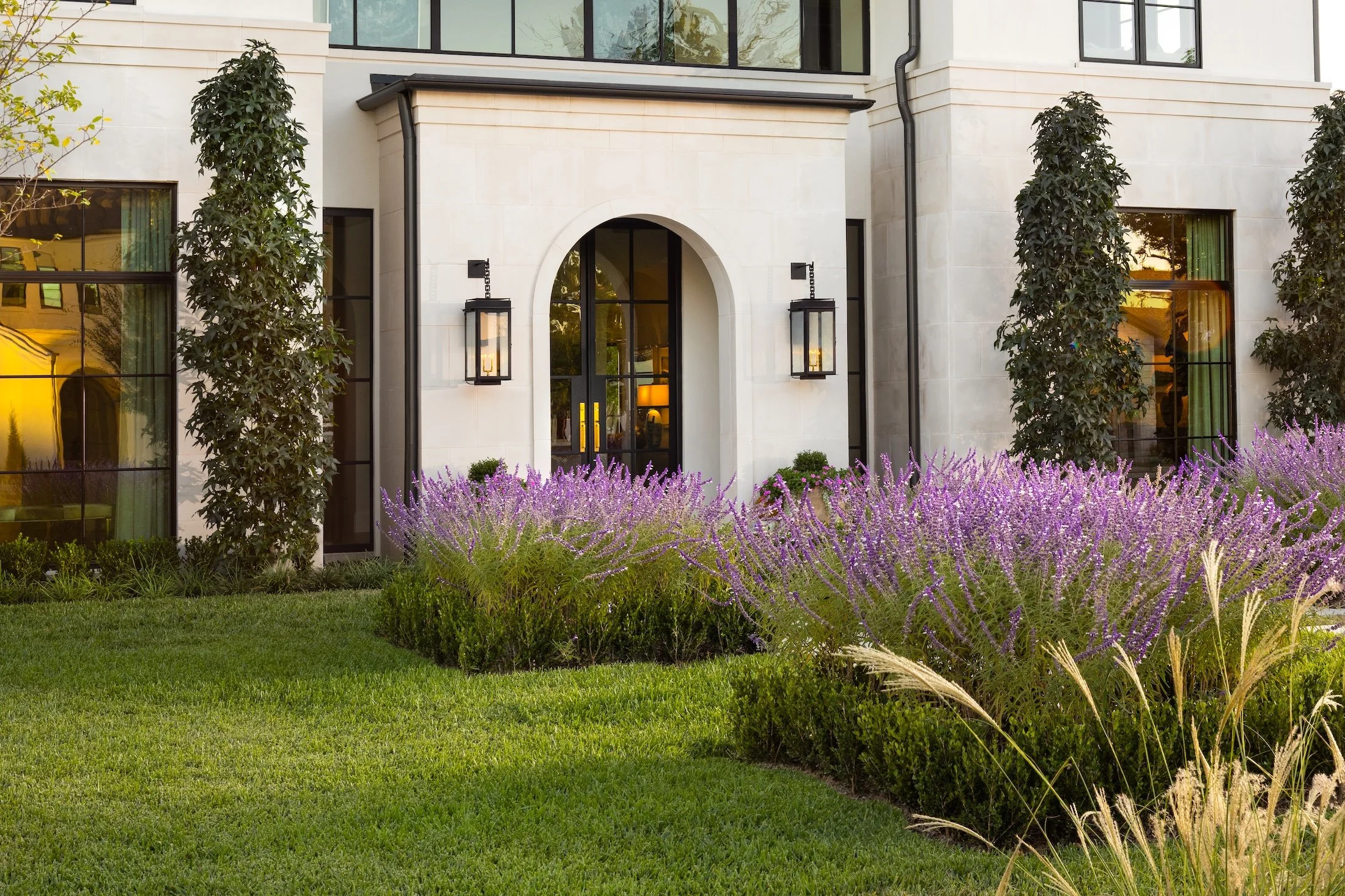 Front of modern house with large windows, white facade, black door, decorative lanterns, green shrubs, and purple flowering plants in the garden.