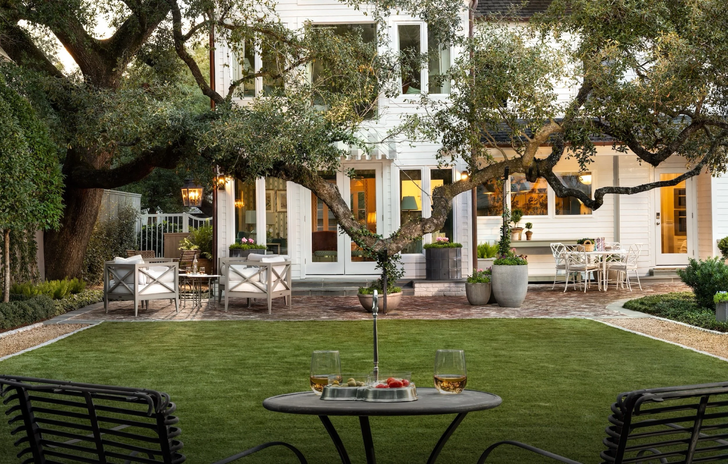 A backyard patio with a large tree, outdoor furniture including a round table with drinks, and a view of the house with multiple windows and potted plants.