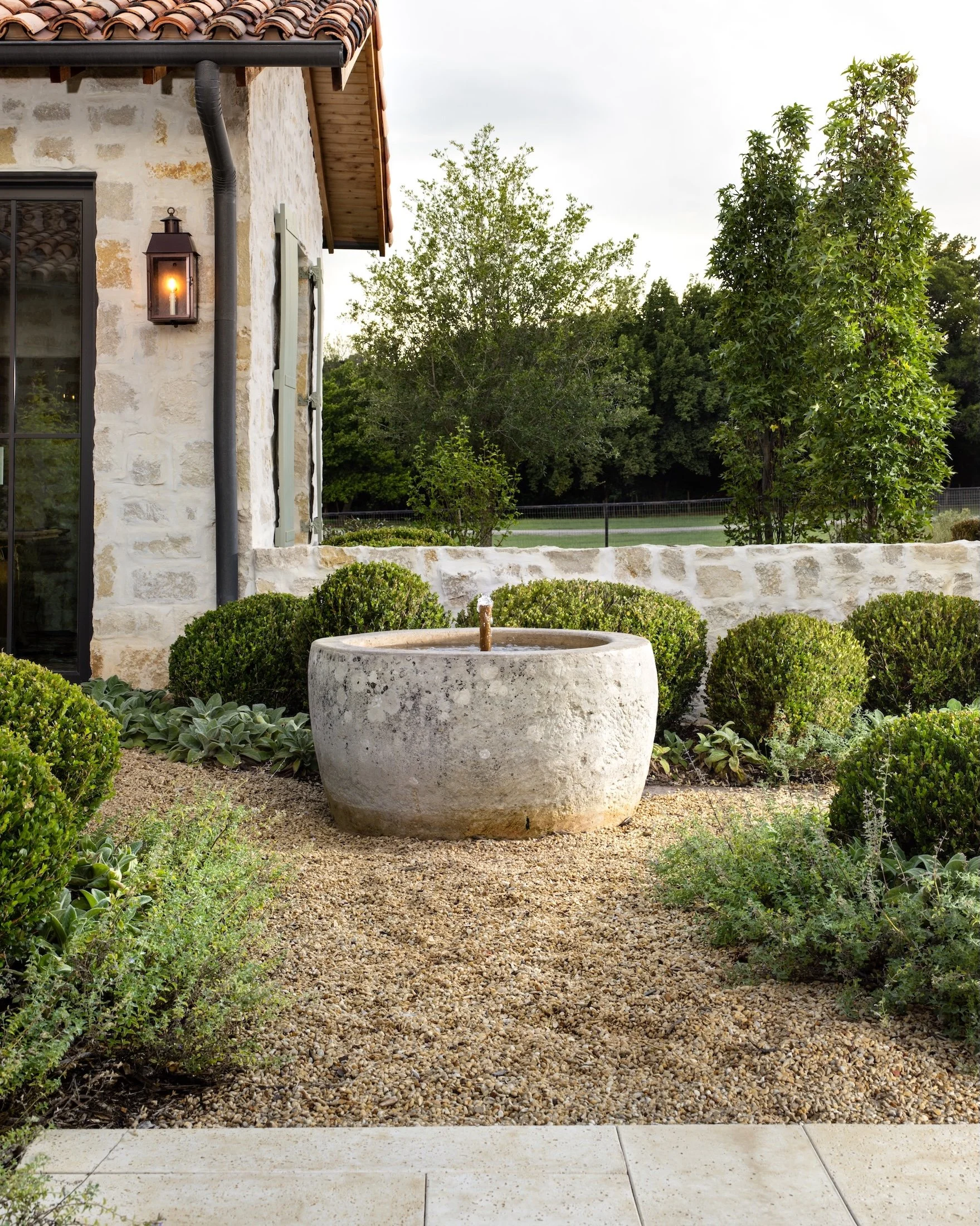 View of a house exterior with stone siding, a black gutter, and a wall-mounted lantern with an illuminated candle-style bulb. In the foreground, there is a large round stone water fountain, surrounded by shrubbery and a pathway with gravel and concrete tiles, with trees and open field in the background.