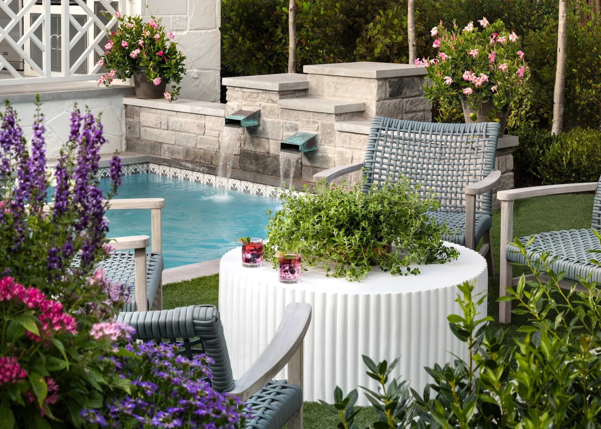 A backyard patio with a pool, pink flowers in pots, plants, and outdoor furniture including chairs and a round table with drinks.