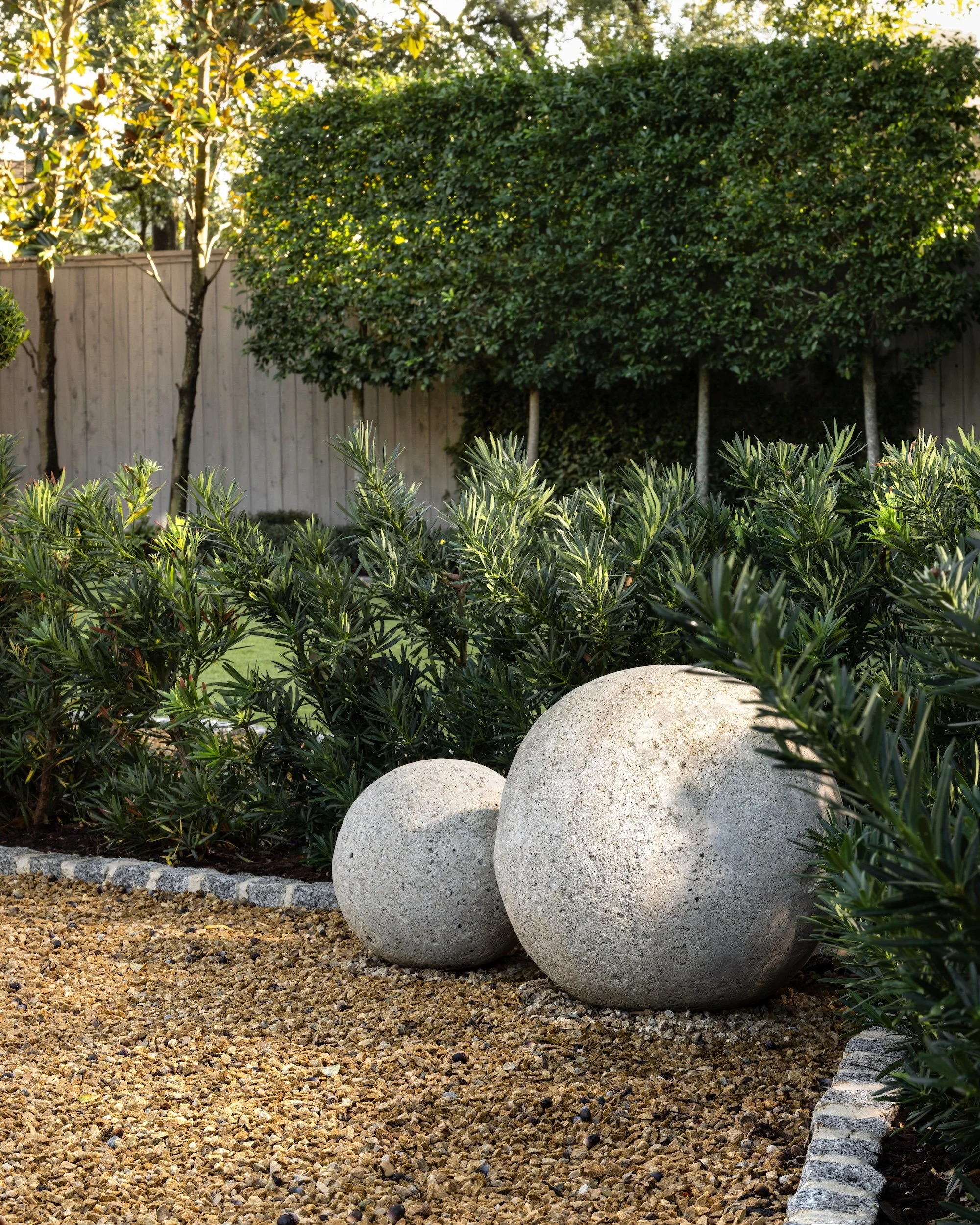 A landscaped yard with large decorative stones, a gravel ground cover, lush green bushes, and trees with dark green leaves, enclosed by a wooden fence.