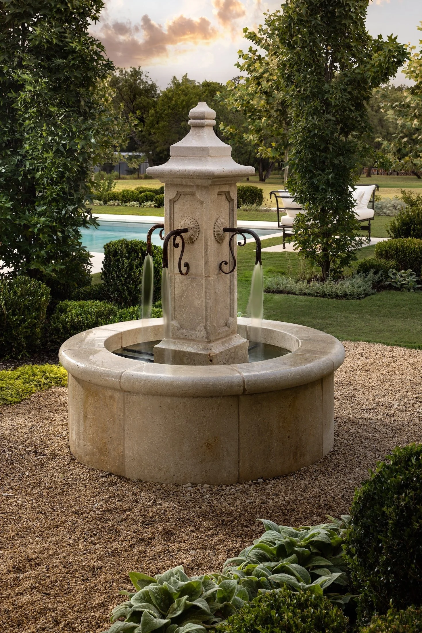 Stone fountain with a circular basin and a tall ornate pillar in a landscaped backyard with shrubs, a swimming pool, and a patio with seating under a cloudy sky.