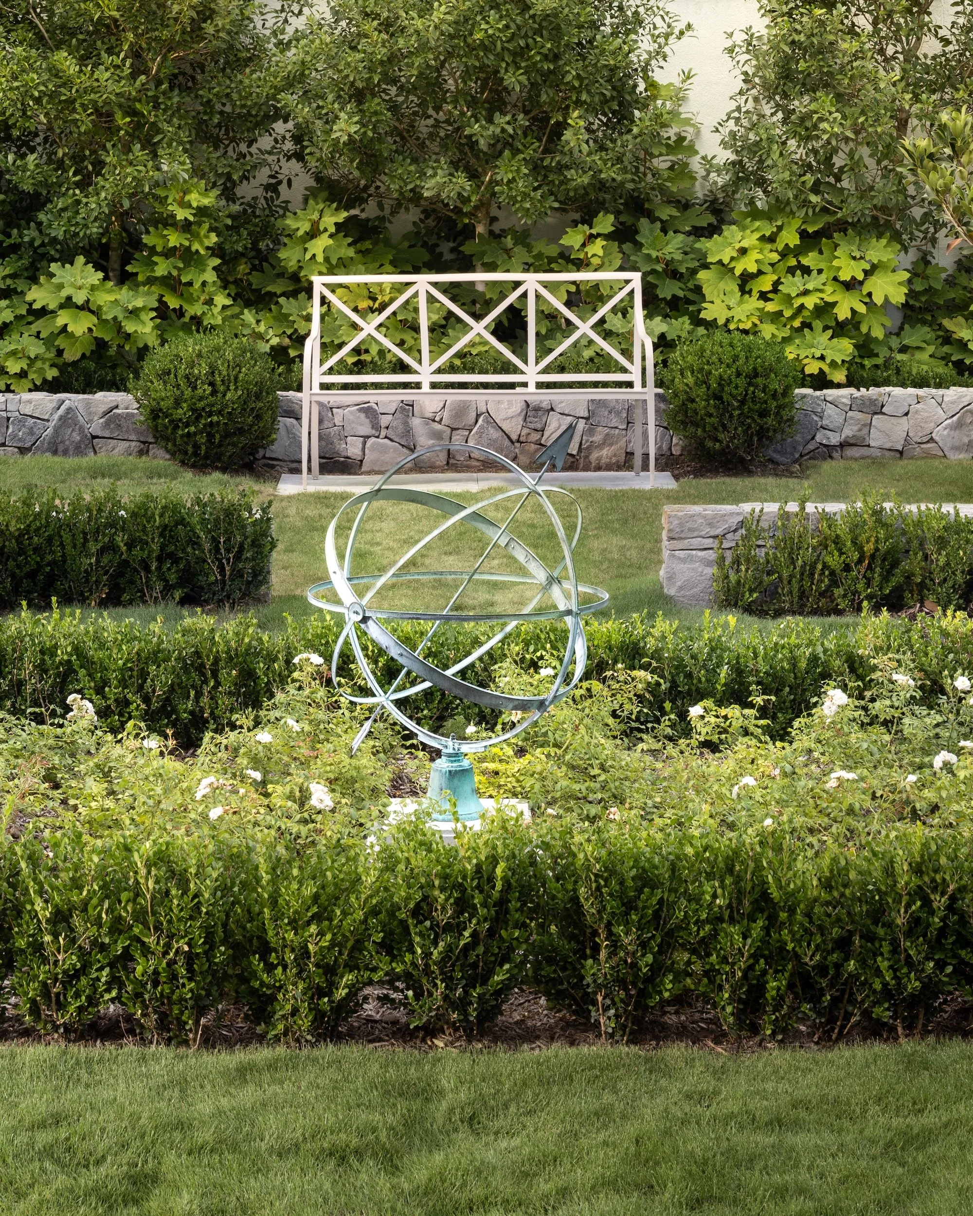A garden with green bushes and trees in the background, a white garden bench, and a metal decorative sculpture in the foreground, all bordered by a stone wall.