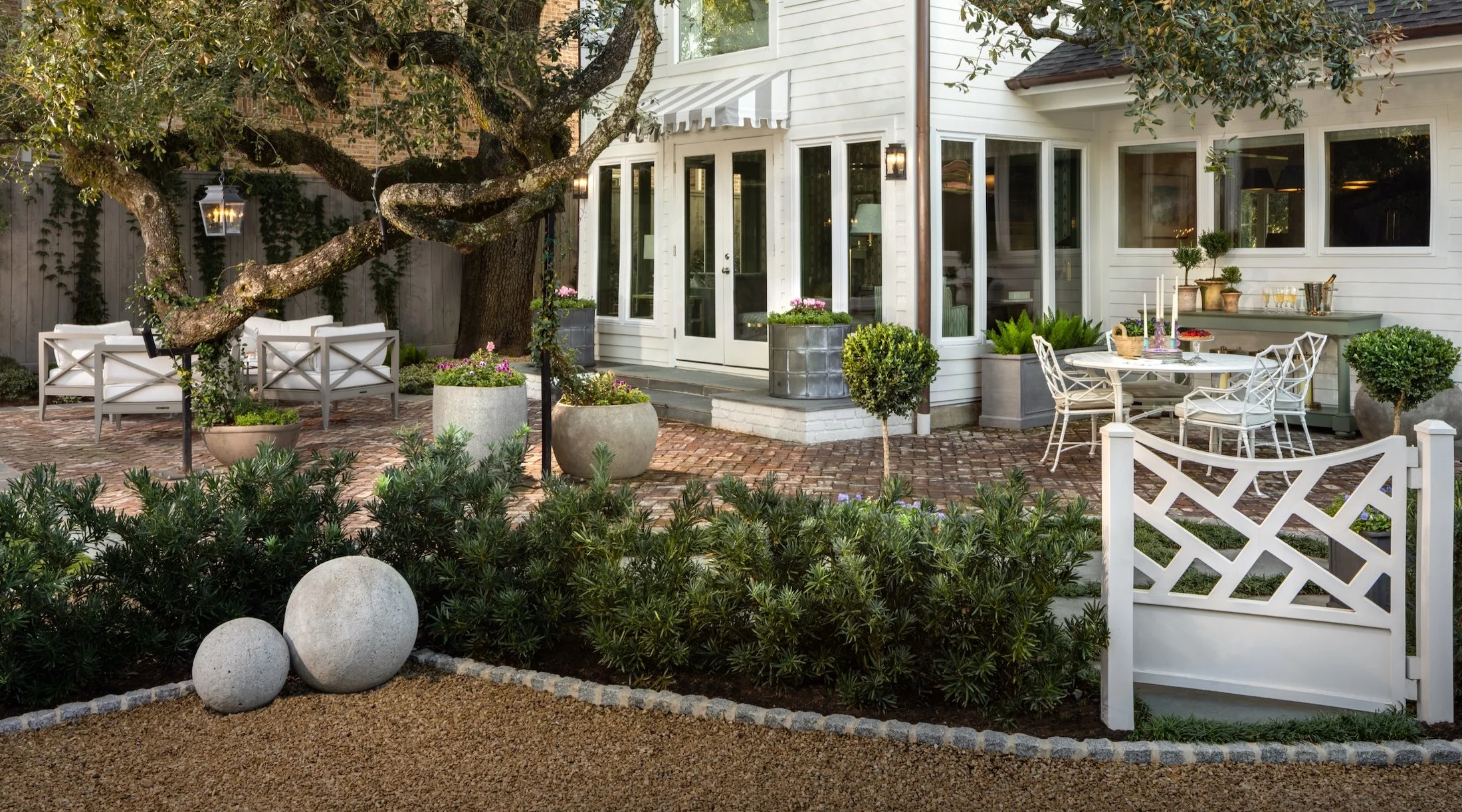 A cozy backyard patio with outdoor furniture, potted plants, and a large tree with sprawling branches. The patio area features a round table with white chairs, a bench, and decorative planters, all set on a brick-paved surface in front of a white house with large windows and a glass door.