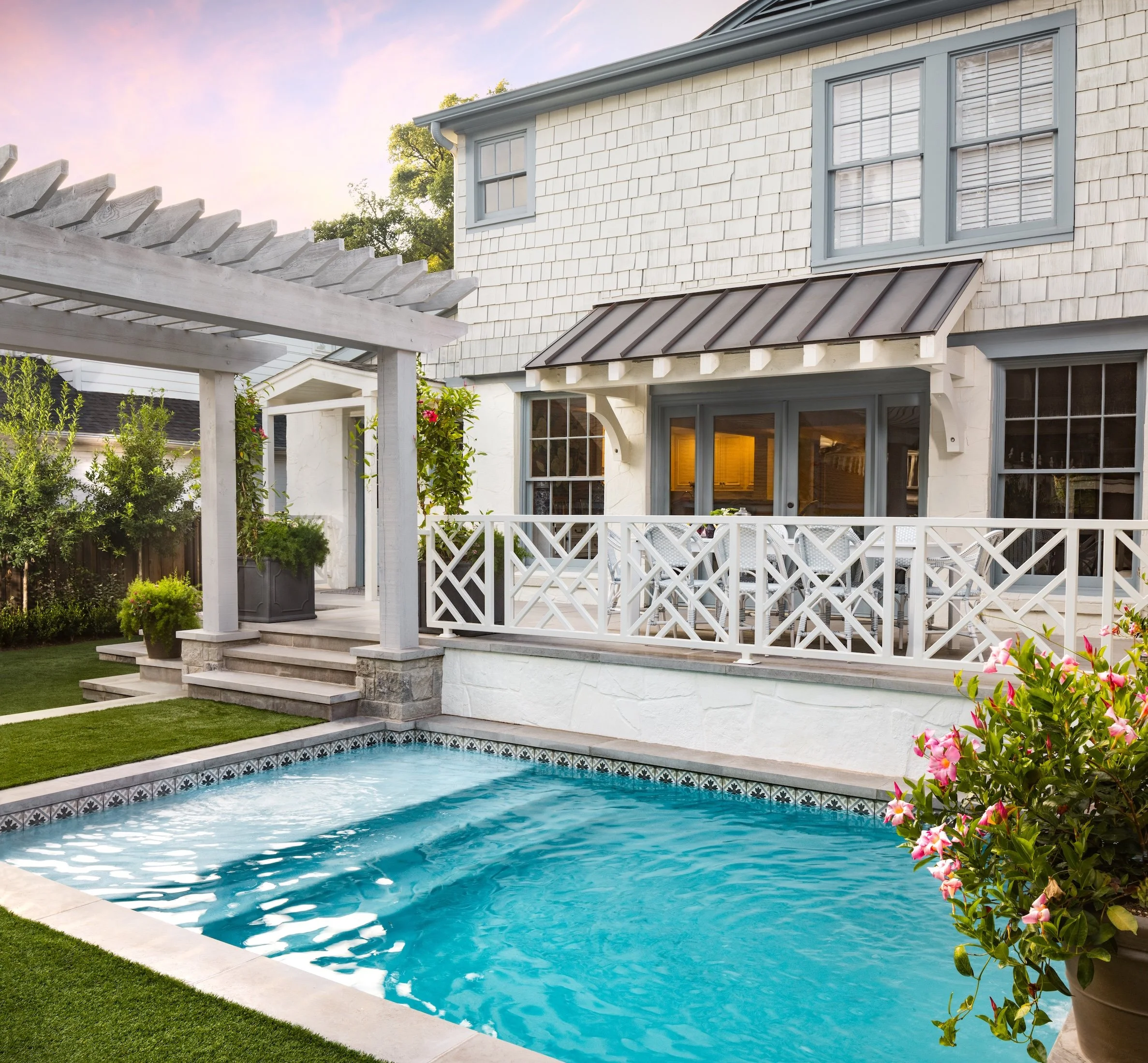 A backyard with a swimming pool, a white house with blue window frames, a small patio, and flowering plants highlighting a sunny, clear day.