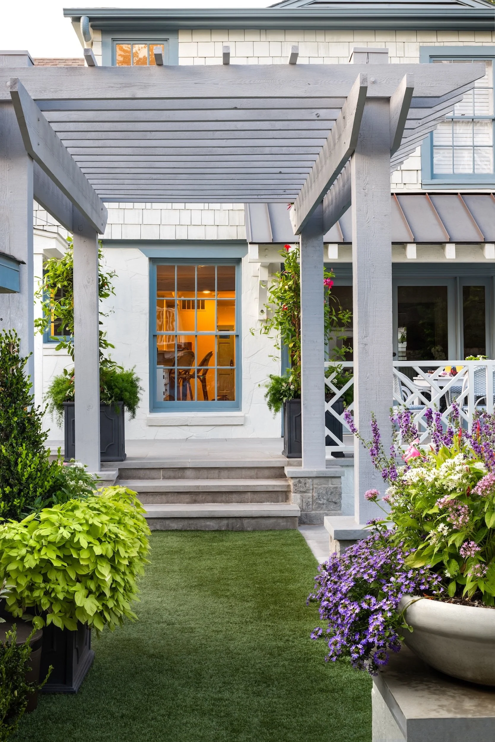 A backyard patio area with a small staircase leading up to a white house with large windows, surrounded by colorful flowers and green plants. There is a pergola structure with white beams overhead and outdoor dining furniture.
