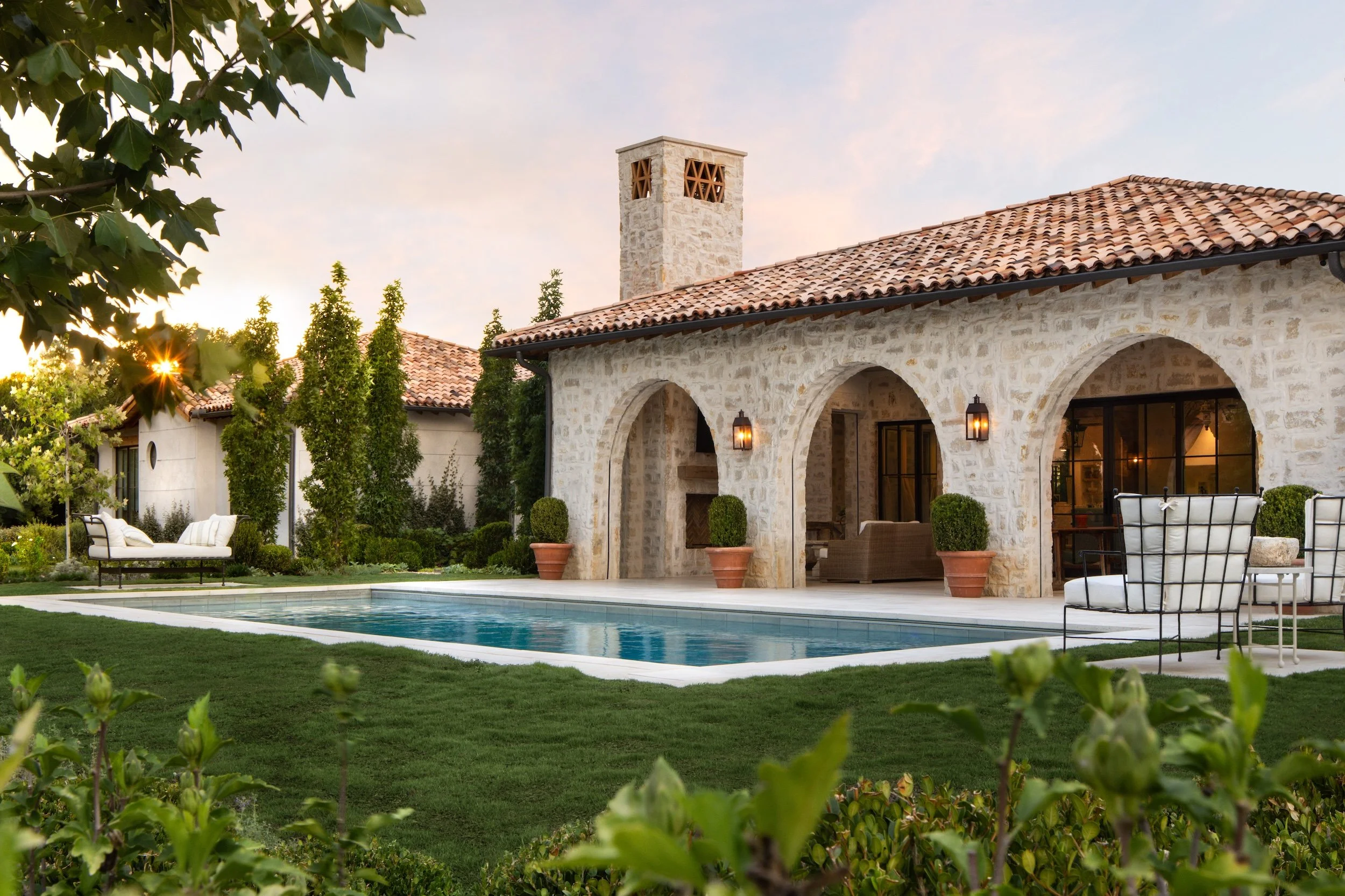 A luxurious house with a stone exterior, red tiled roof, and a chimney, surrounded by a garden with a swimming pool and outdoor seating area, during sunset.