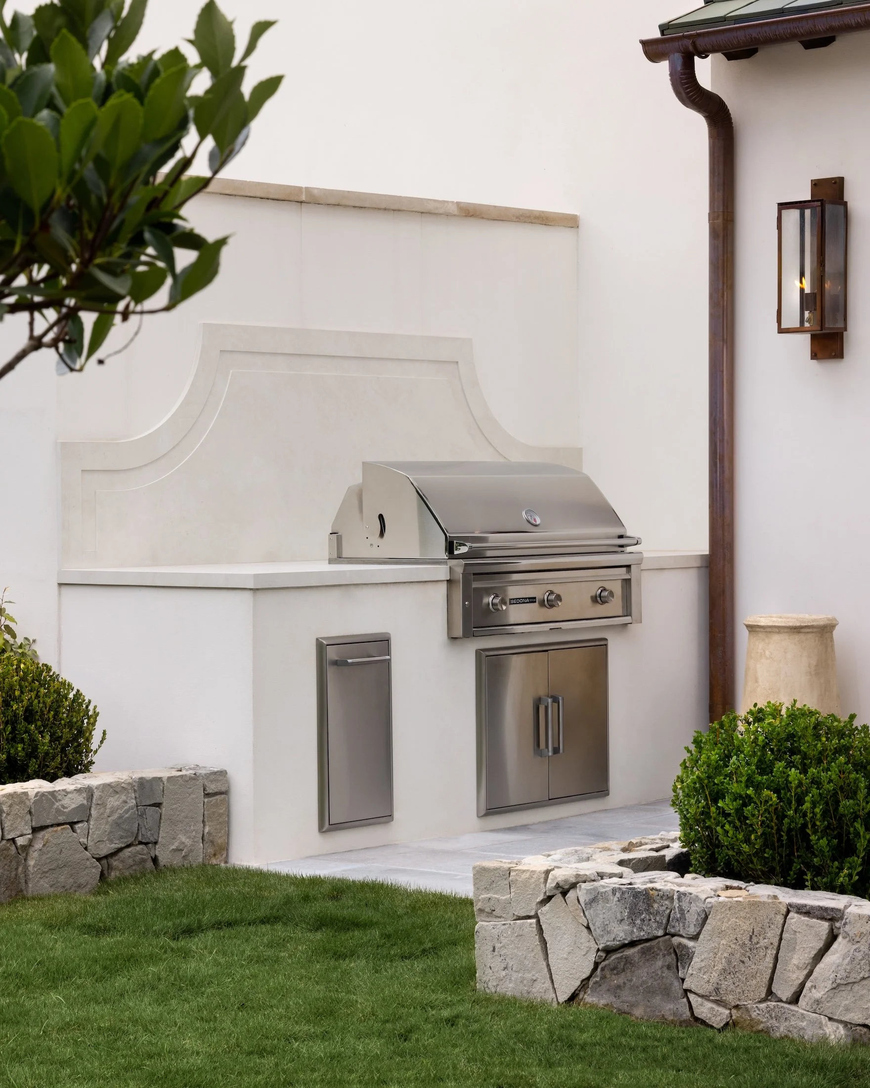An outdoor kitchen area with a stainless steel grill, a stone wall, green bushes, and a white stucco wall with a lantern and a drain pipe.