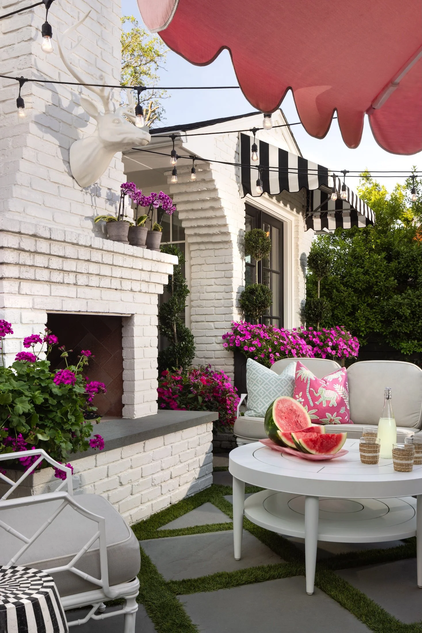 A cozy outdoor patio featuring a white brick fireplace with a mounted white deer head sculpture, potted purple orchids, and vibrant pink petunias. There are string lights, a pink watermelon, a carafe of water, and seating with patterned pillows under black-and-white striped awnings.