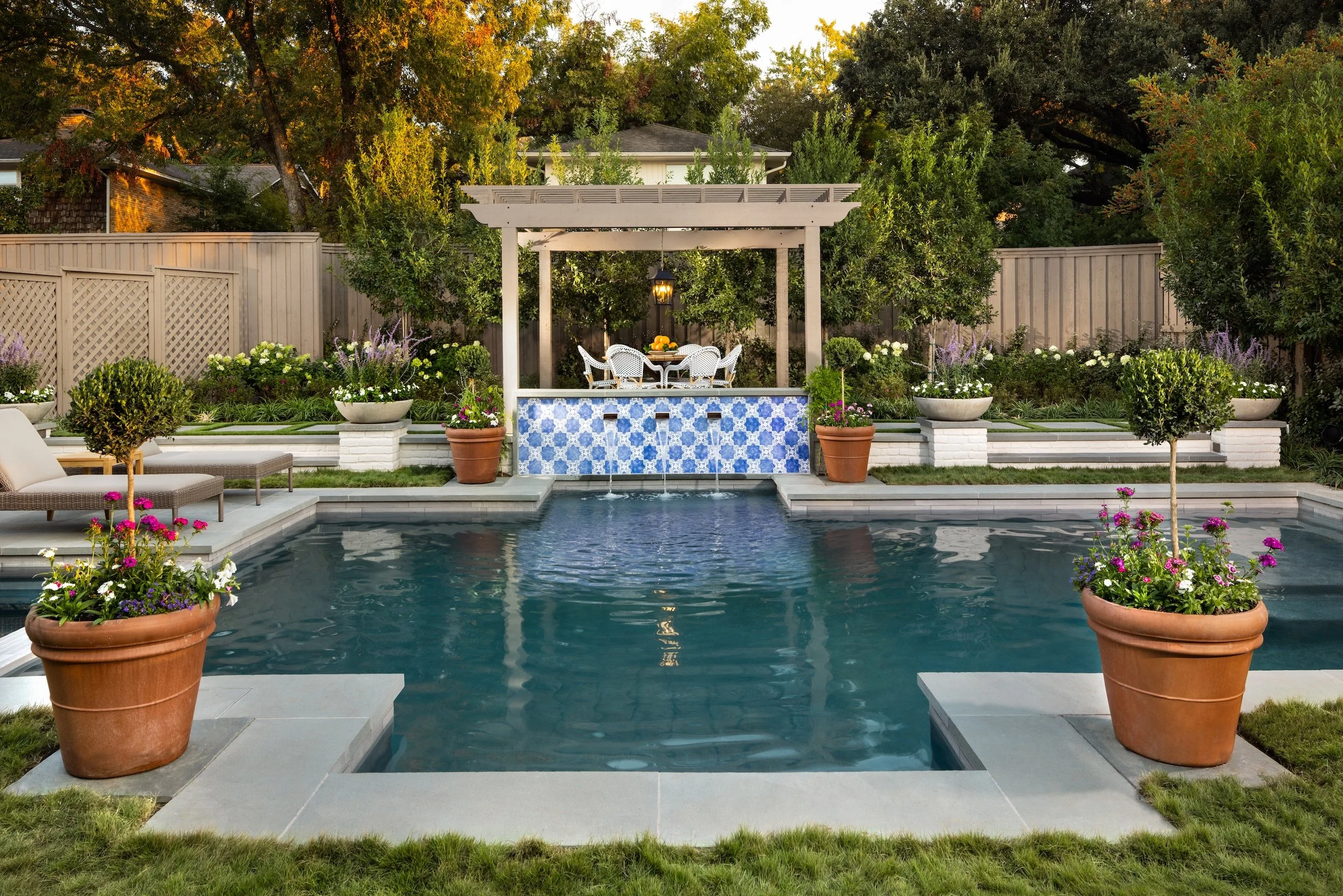 A backyard swimming pool with a surrounding patio and lush greenery, including potted plants and trees. There is a white pergola with outdoor dining furniture and a small fountain in the middle of the scene, illuminated by a hanging lantern, during evening time.