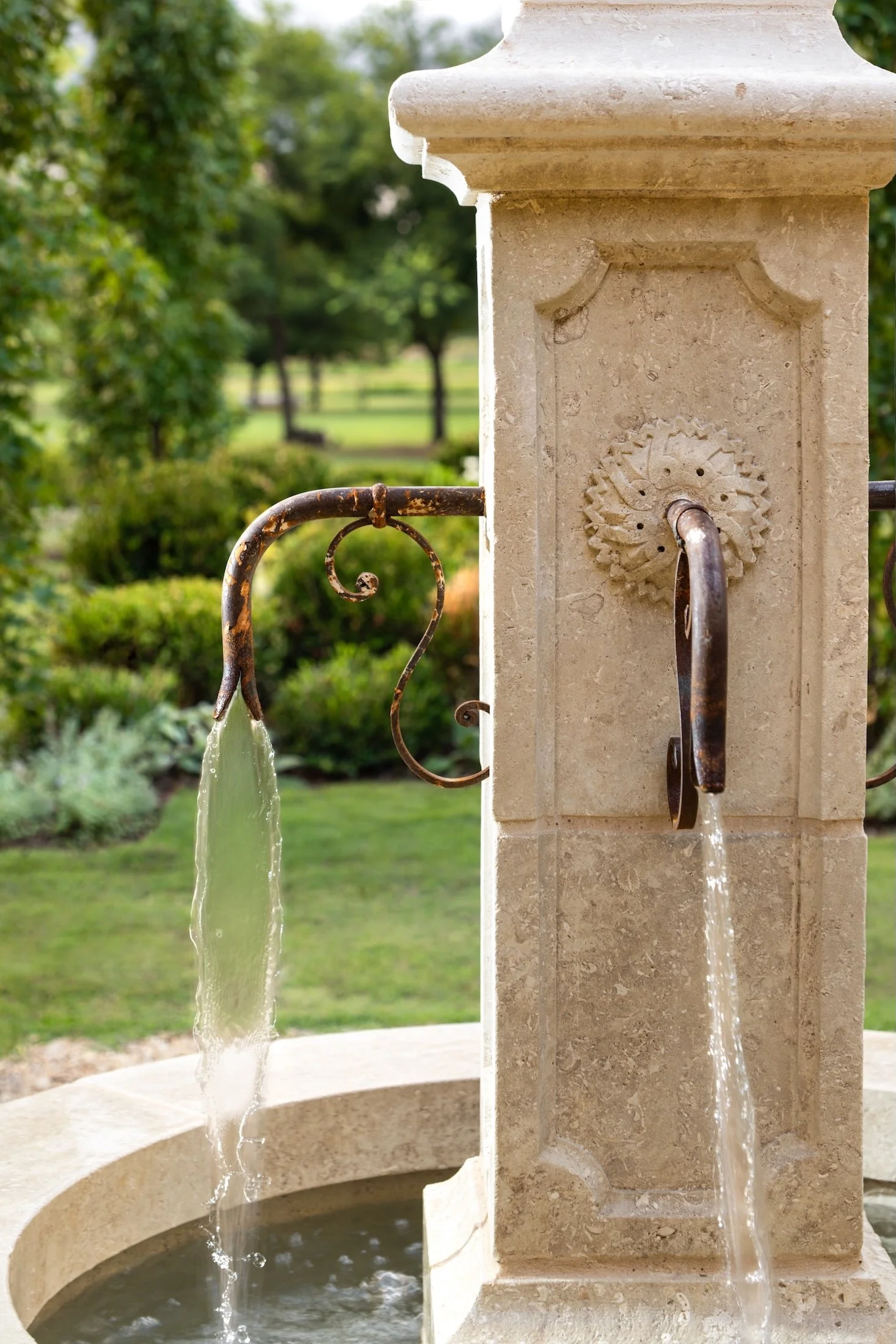 Two water fountains built into a stone pillar with metal spouts, water flowing from both, set against a background of lush green trees and shrubs.