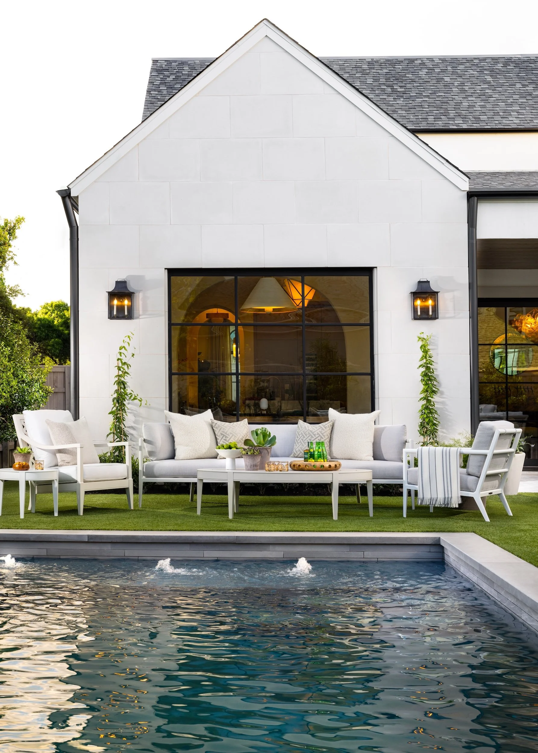 A backyard scene with a white house featuring large black-framed windows, outdoor seating with beige cushions, a table with green bottles, and a swimming pool with rippling water in the foreground.