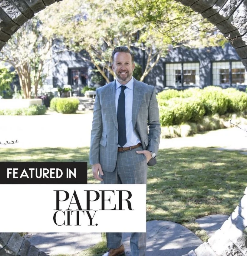 A man in a gray plaid suit smiling outdoors under a stone arch. A banner in the lower left says "Featured in Paper City." The background shows trees, bushes, and a black building with windows.