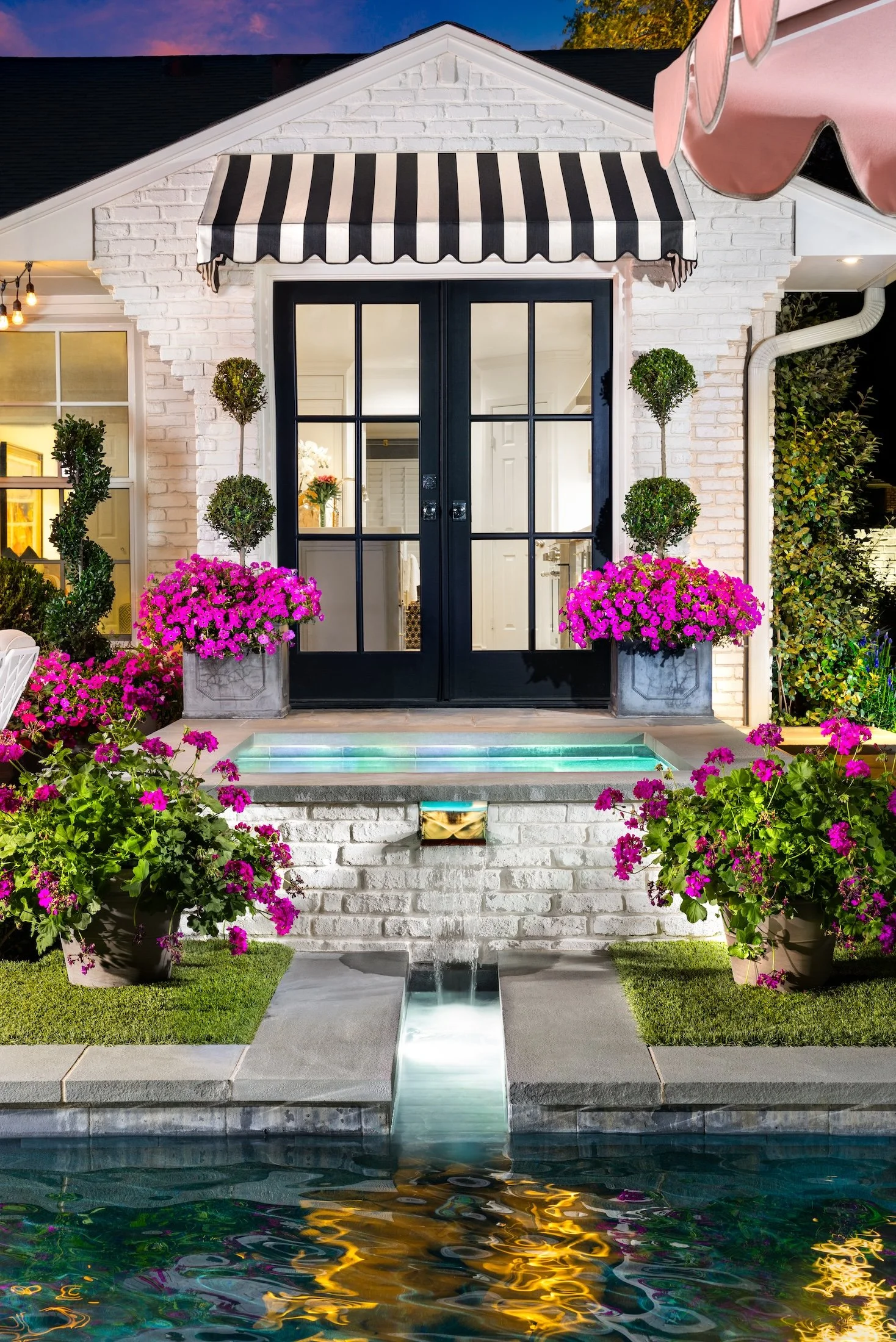 A stylish house with a white brick exterior, black double doors with glass panes, and a black-and-white striped awning. The front yard features vibrant pink flowers, potted topiary trees, and a small illuminated water feature leading to a pool, all illuminated at dusk.