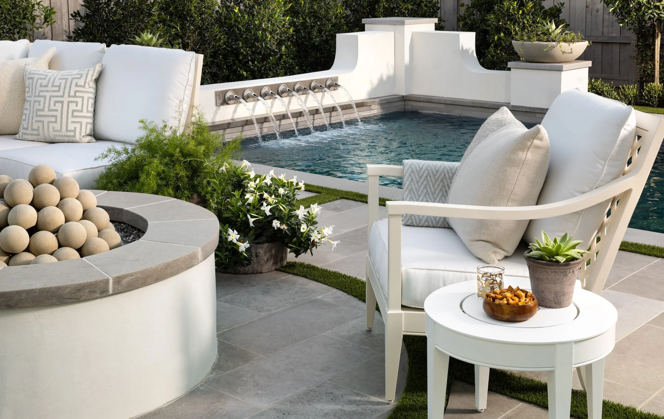 Outdoor patio with white cushioned chairs, a small round table with snacks, a fire pit with beige spheres, and a pool with an integrated waterfall feature surrounded by lush greenery.