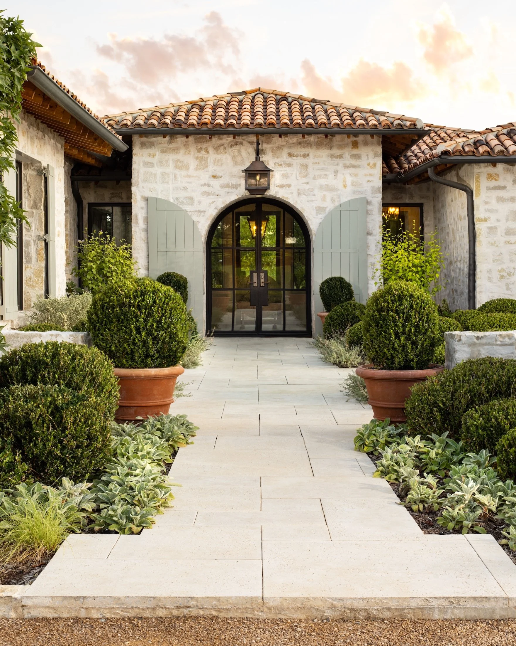 A stone house with a tile roof and green shutters, featuring a gated arched doorway, surrounded by potted plants and shrubs, with a cloudy sky in the background.