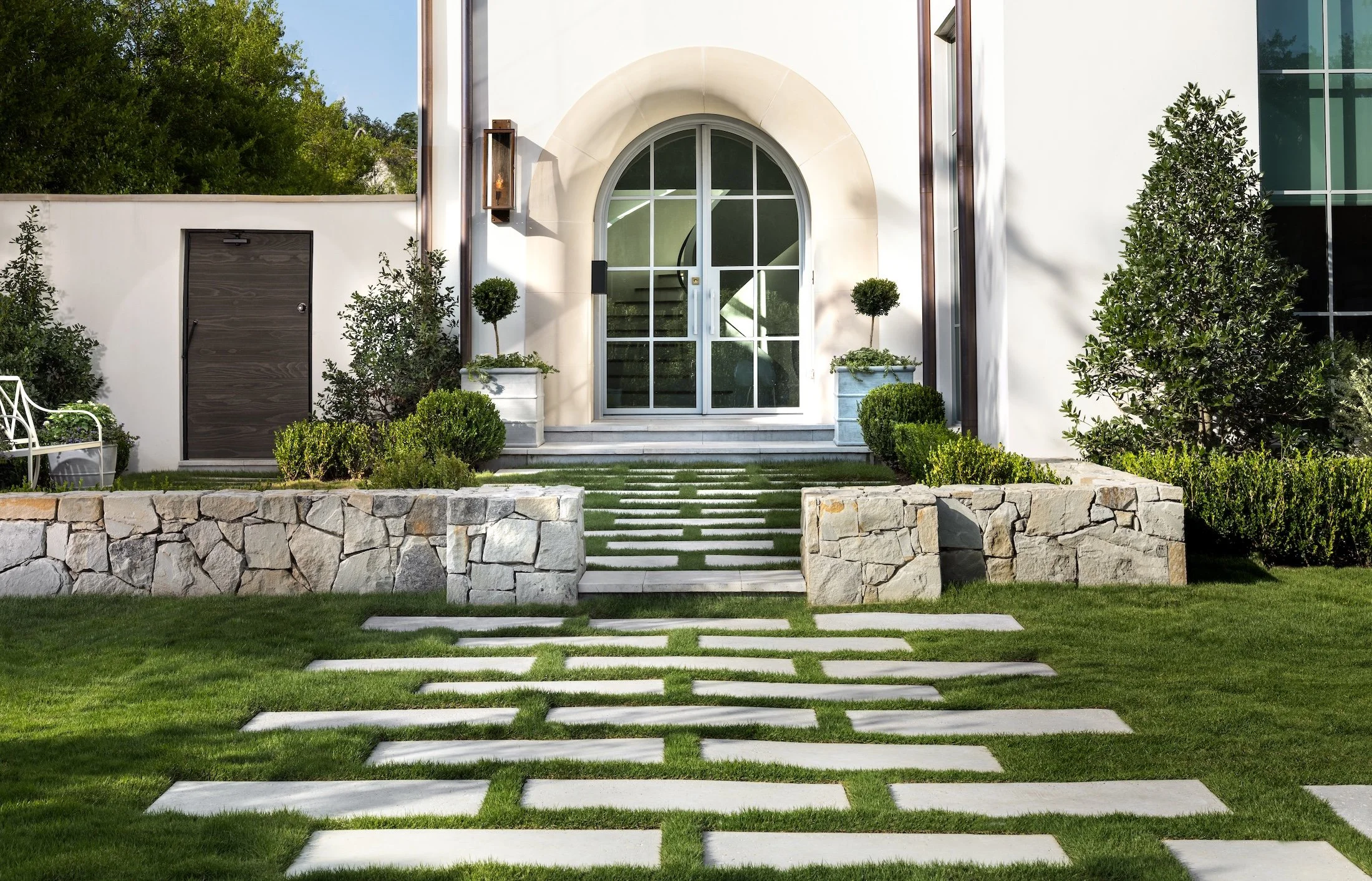Front entrance of a modern house with large glass doors, stone steps, green lawn, and potted plants.