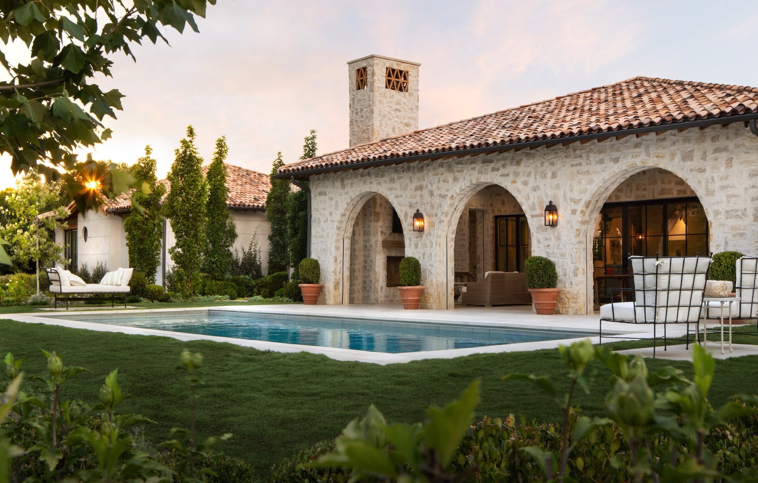 A backyard house with a swimming pool and outdoor furniture, surrounded by greenery and trees, during sunset.