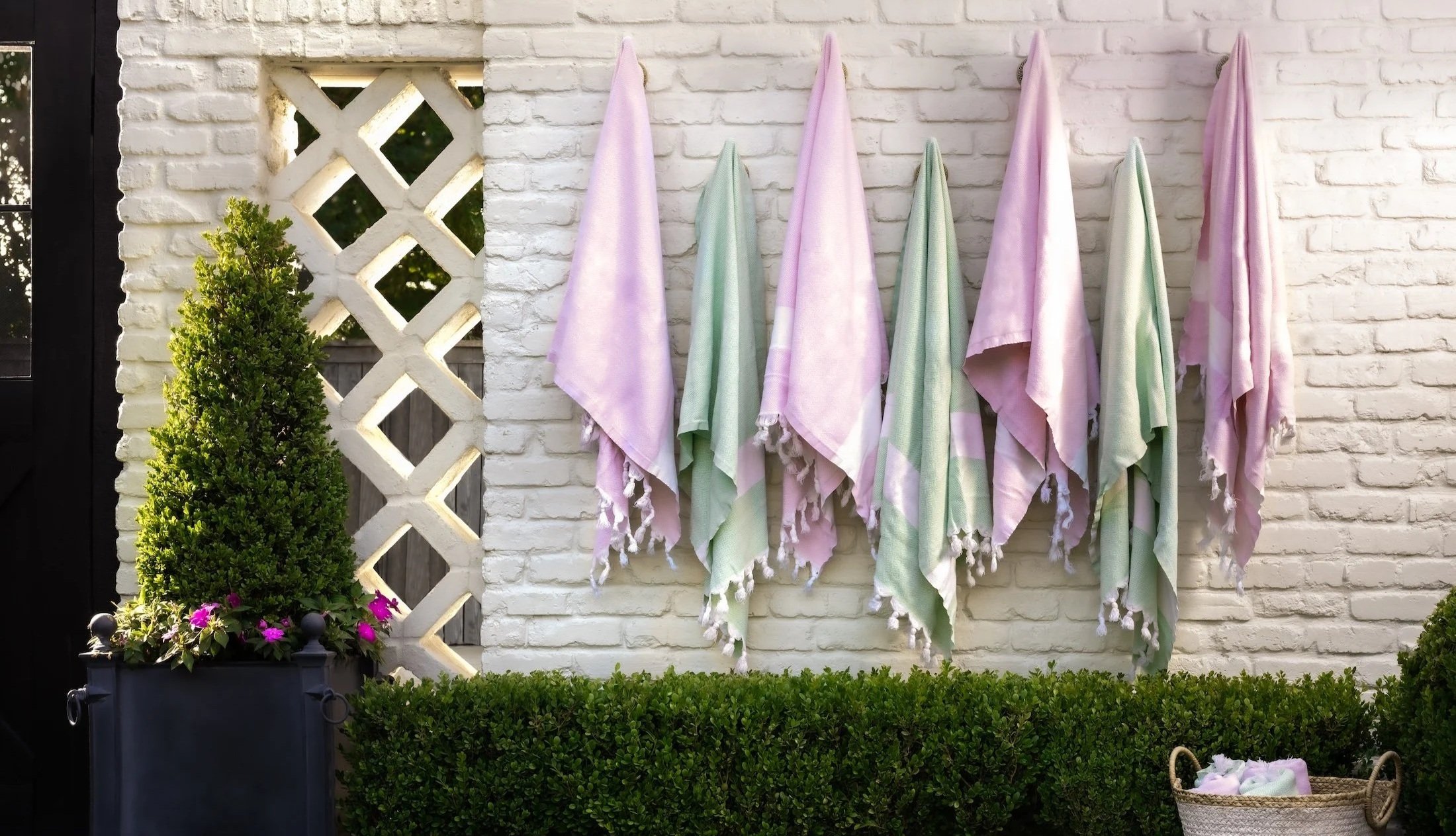 Outdoor scene with pastel-colored towels hanging on a white brick wall, green shrubbery and pink flowers in a black planter, decorative lattice, and a basket with laundry on the ground.
