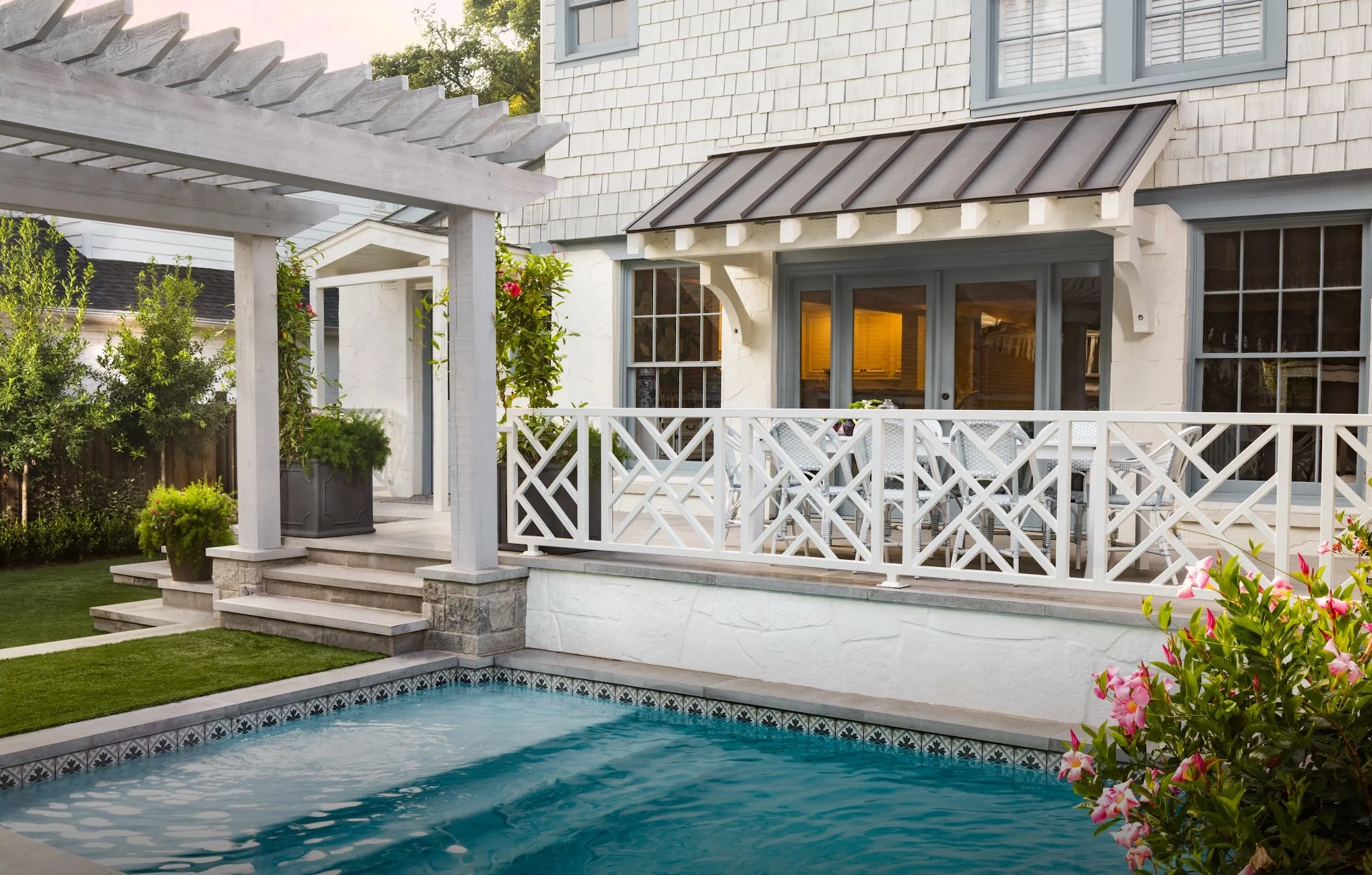 Backyard view featuring a swimming pool, white patio with steps, white wooden fencing, and house with gray siding, large windows, and a metal roof over the door, surrounded by greenery and flowering plants.