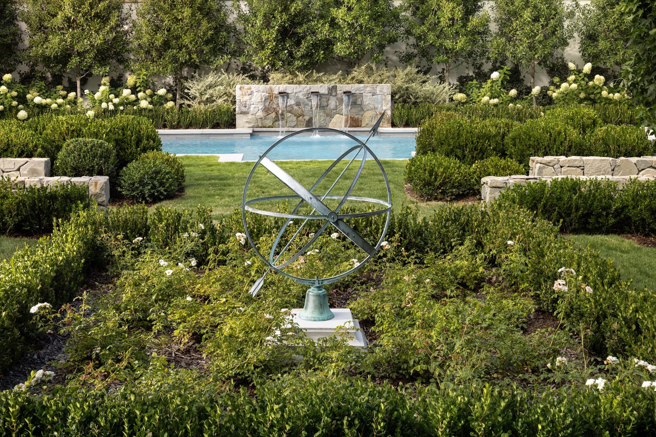 Landscape view of a lush garden featuring a water fountain with three streams of water, green bushes, and white flowering plants, with tall green trees in the background.