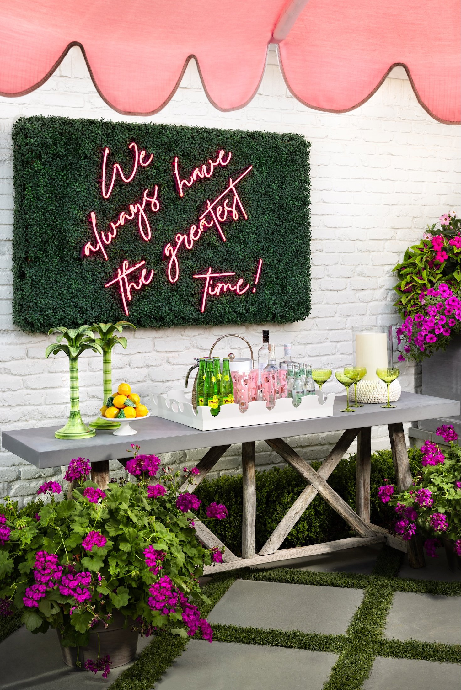 Outdoor party setup with a pink banner, a green wall with neon pink sign reading 'We always have the greatest time!', a table with green glasses, bottled drinks, a fruit bowl, pink cups, and candles, surrounded by pink and purple flowers and greenery.