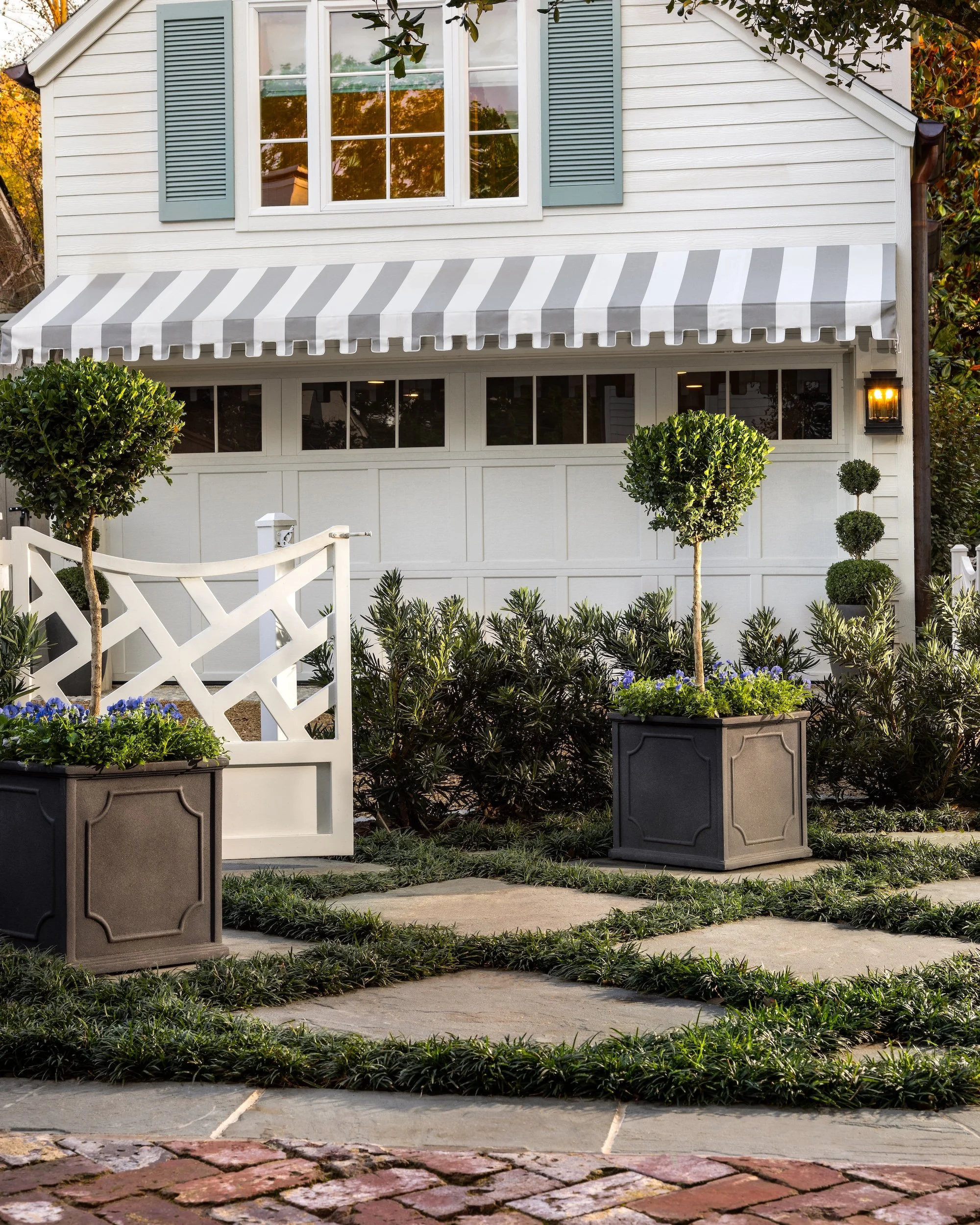 Front yard of a house with a white facade, a gray and white striped awning, decorated potted plants, manicured bushes, and a brick walkway.