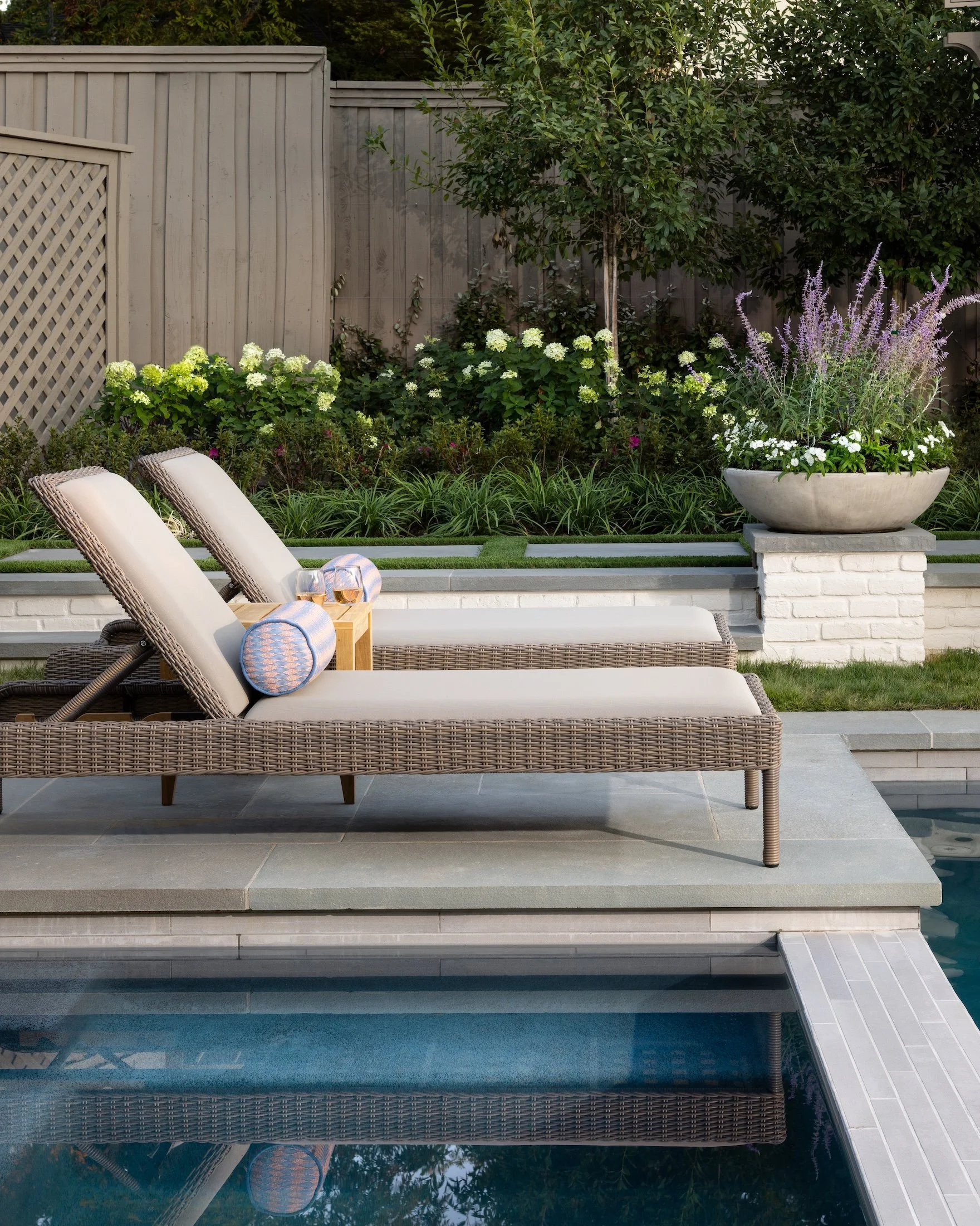 A backyard scene with a swimming pool, a white lounge chair with a blue towel, a large decorative planter with purple and white flowers, and a wooden fence with green trees and bushes in the background.