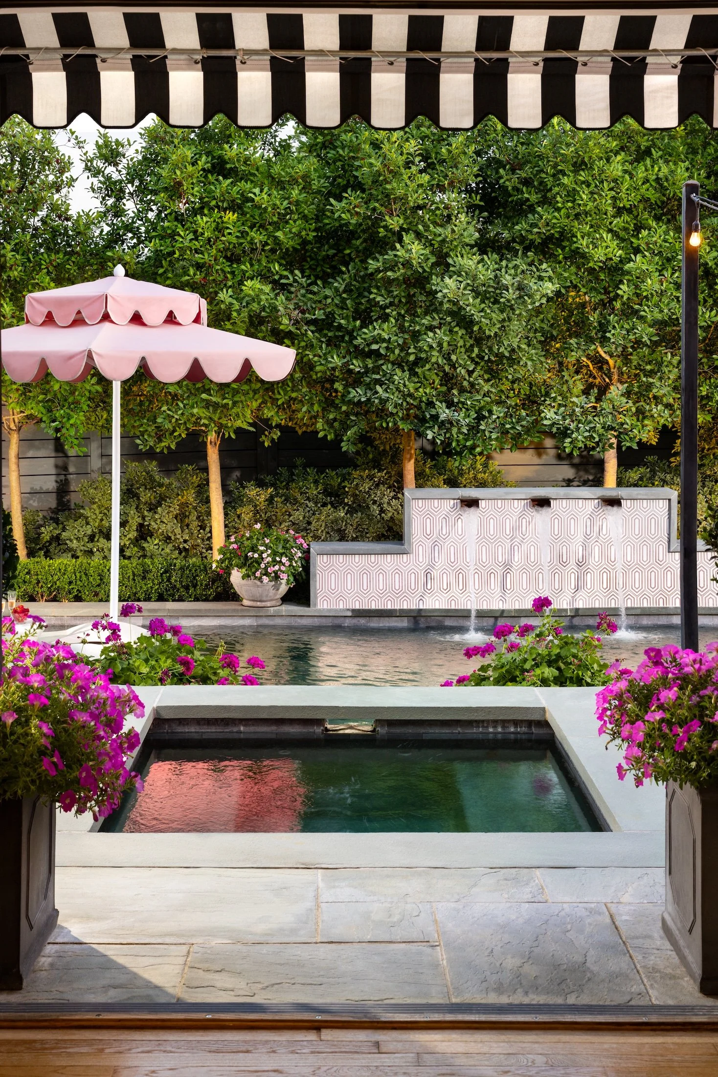 View of a backyard with a small pool, pink flowers in pots, a pink patio umbrella, green trees, and a black and white striped awning.