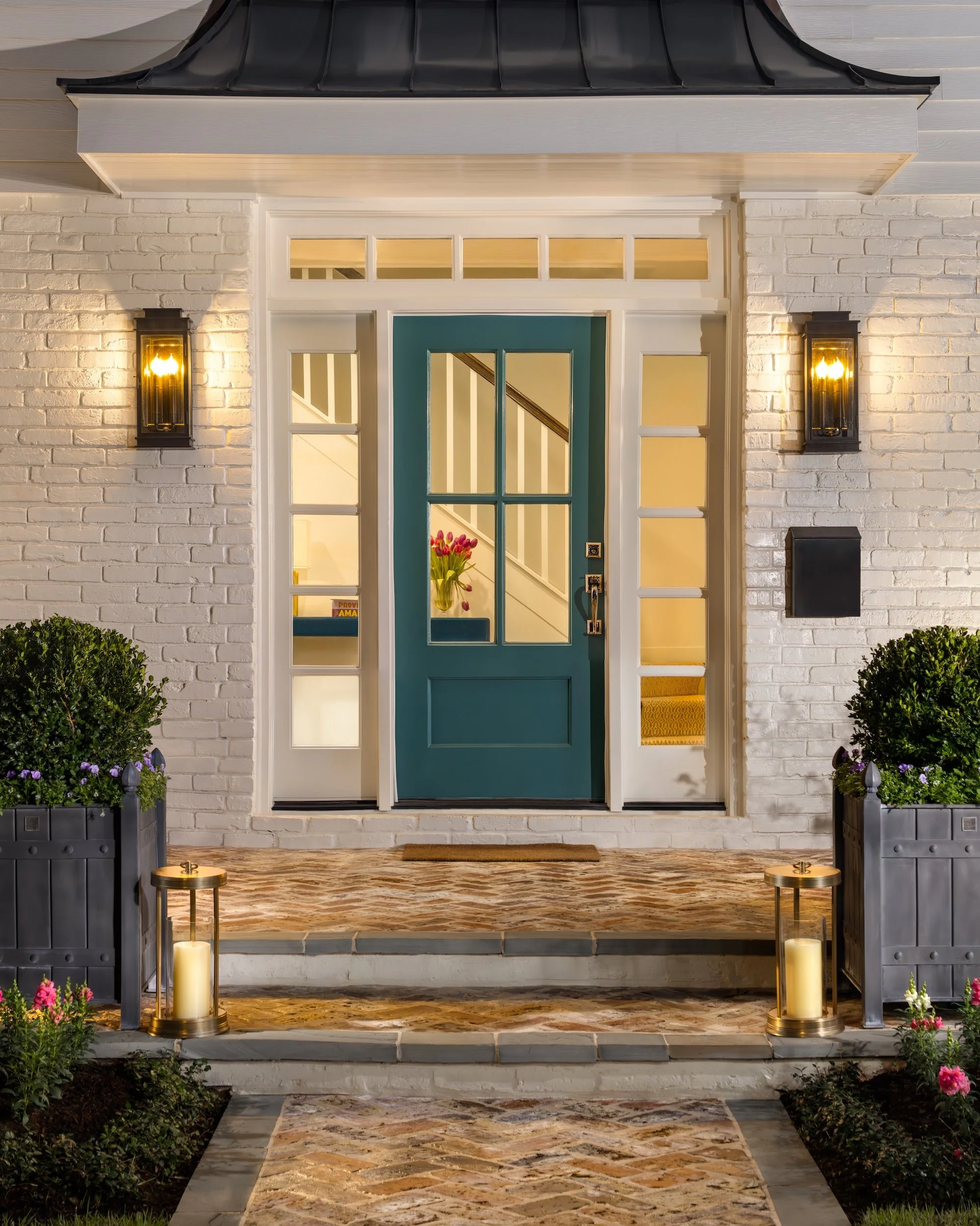 Front porch of a house with a teal door, white brick wall, black wall-mounted lanterns, potted bushes, candles, and a stone pathway.