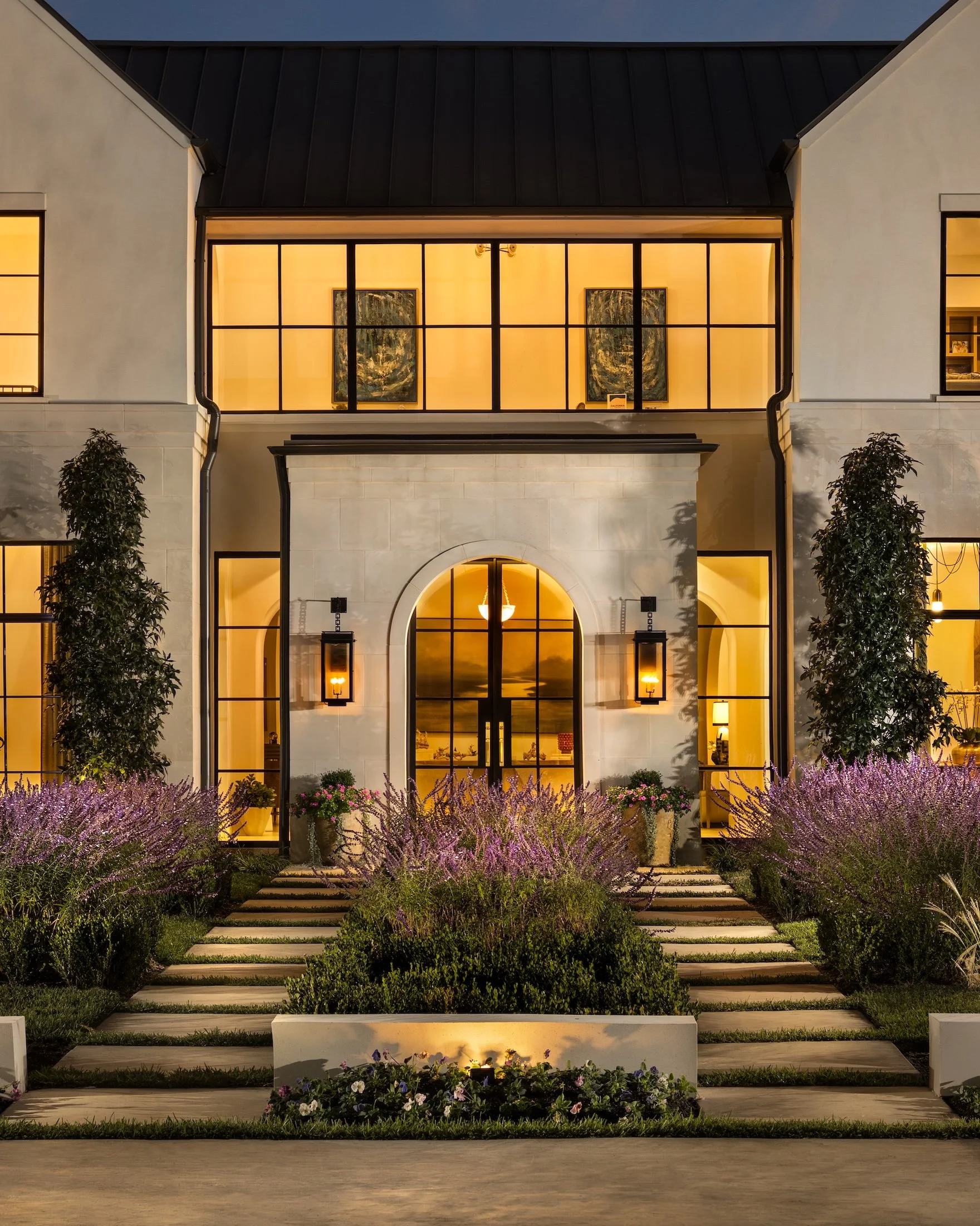A modern house with large illuminated windows at twilight, landscaped with purple and pink flowers, bushes, and a pathway leading to the front entrance.
