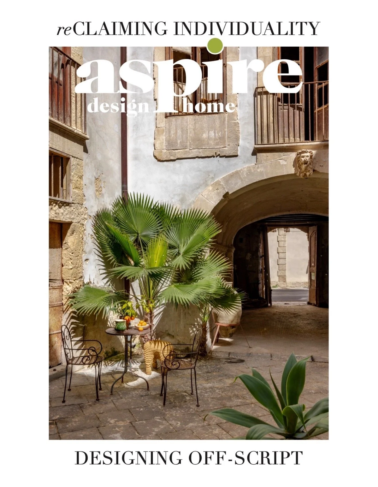 an outdoor courtyard with stone paving, green plants, a small round metal table with chairs, and a building with stone and plaster walls, an archway, and balconies with railing