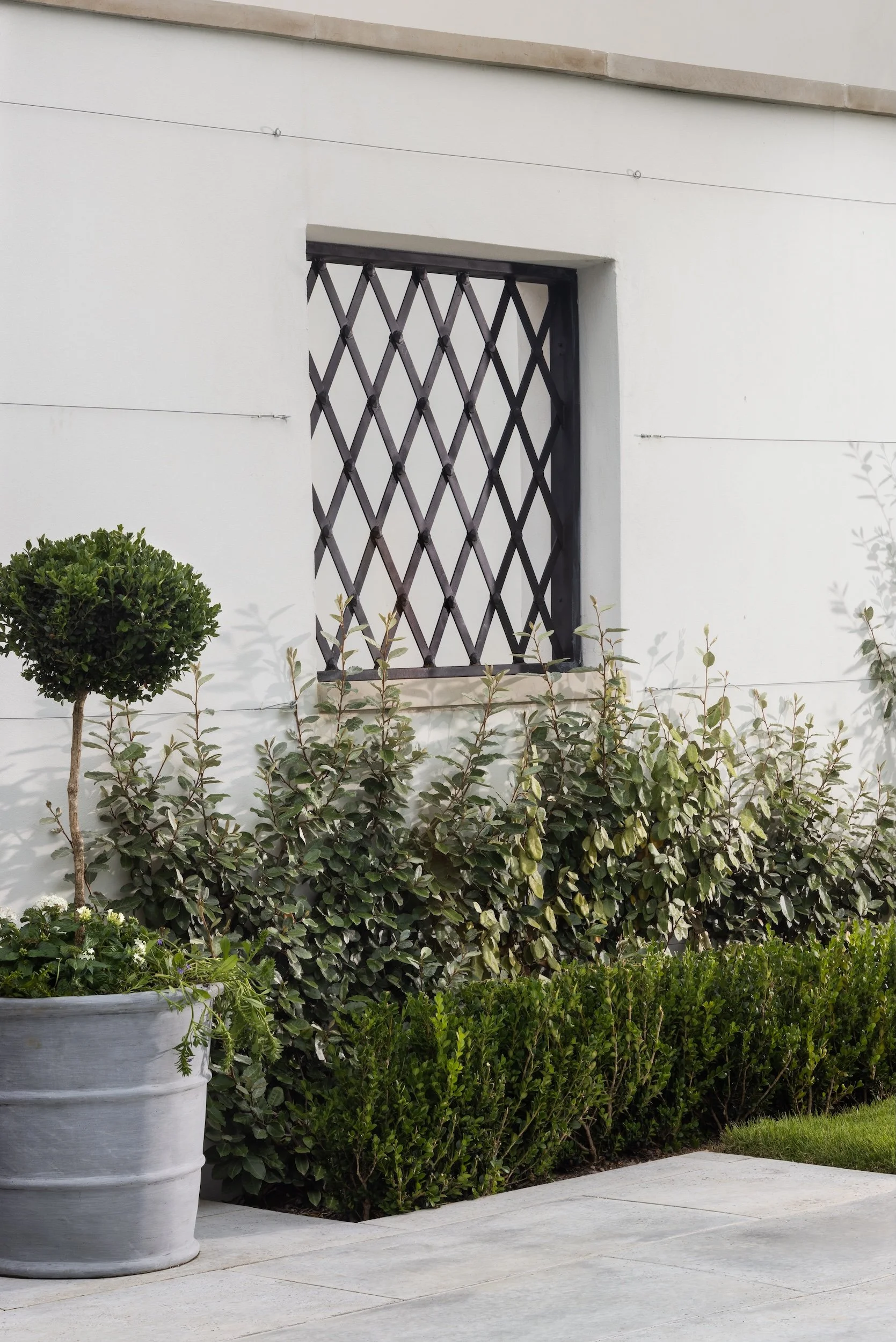 A white exterior wall with a decorative black metal window grate, surrounded by well-maintained green bushes and a large gray planter on a concrete sidewalk.