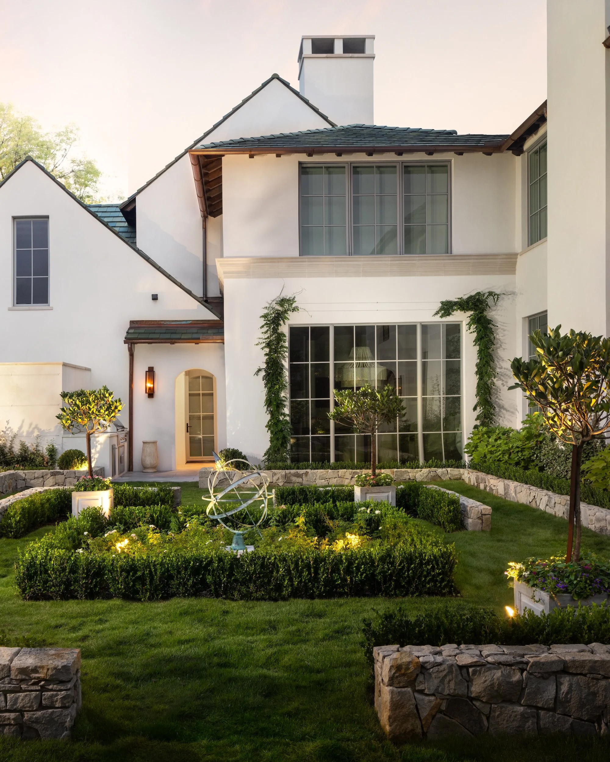 Front view of a modern white house with large windows, a garden with trimmed bushes, small trees, outdoor lights, and a decorative metallic armillary sphere sculpture in the yard. The house features a sloped roof with dark shingles and a small balcony area.